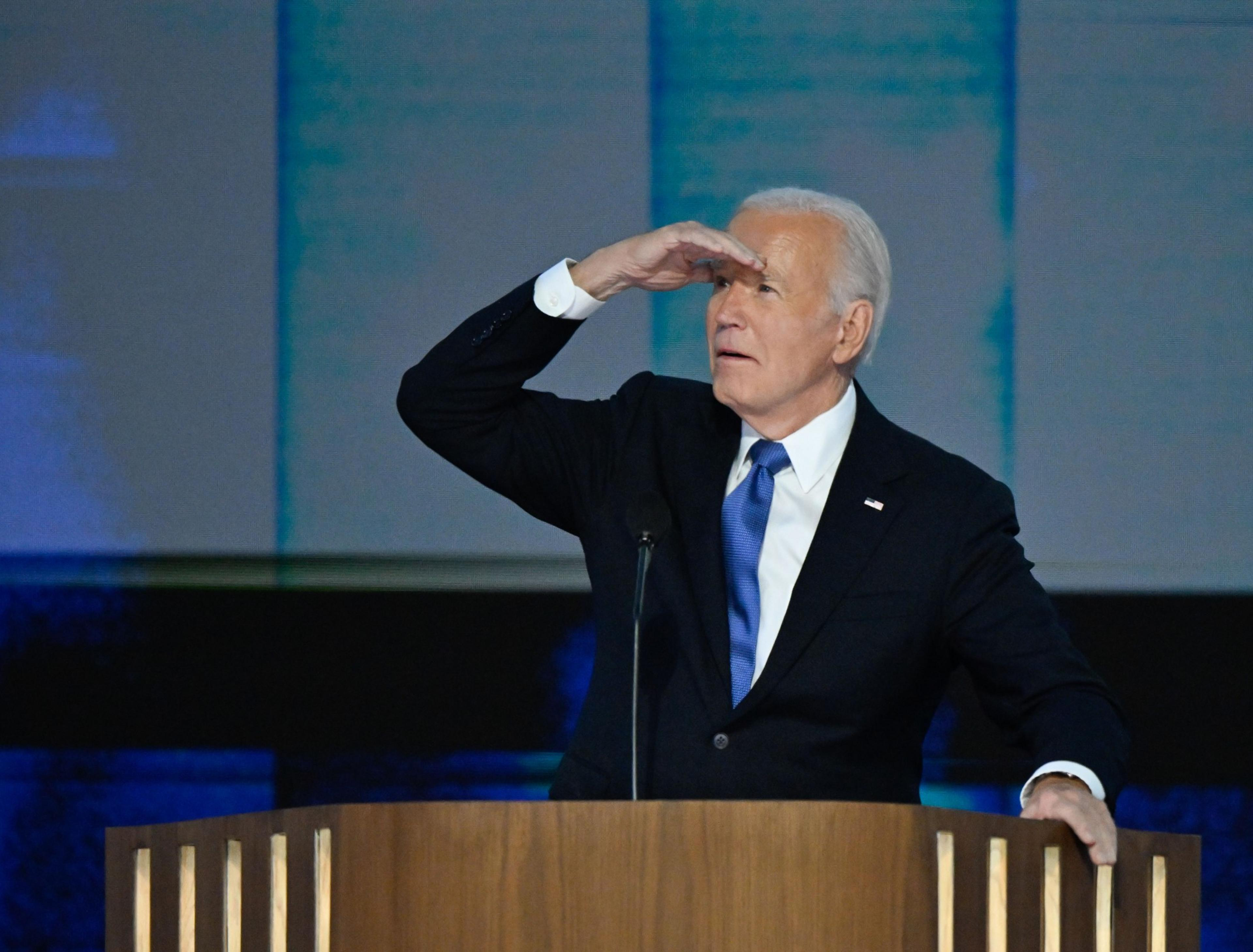 A man in a suit stands at a podium, shielding his eyes with his hand as if looking into the distance. The background is a soft blue with vertical lines.