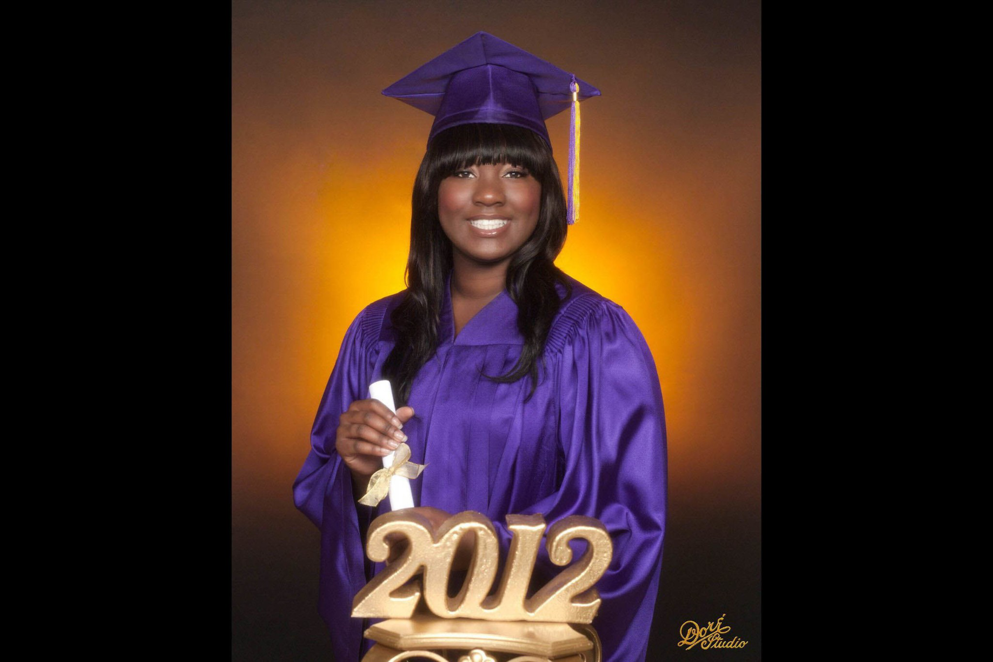 A smiling woman in a purple graduation gown and cap holds a diploma with a 2012 gold decoration in front.