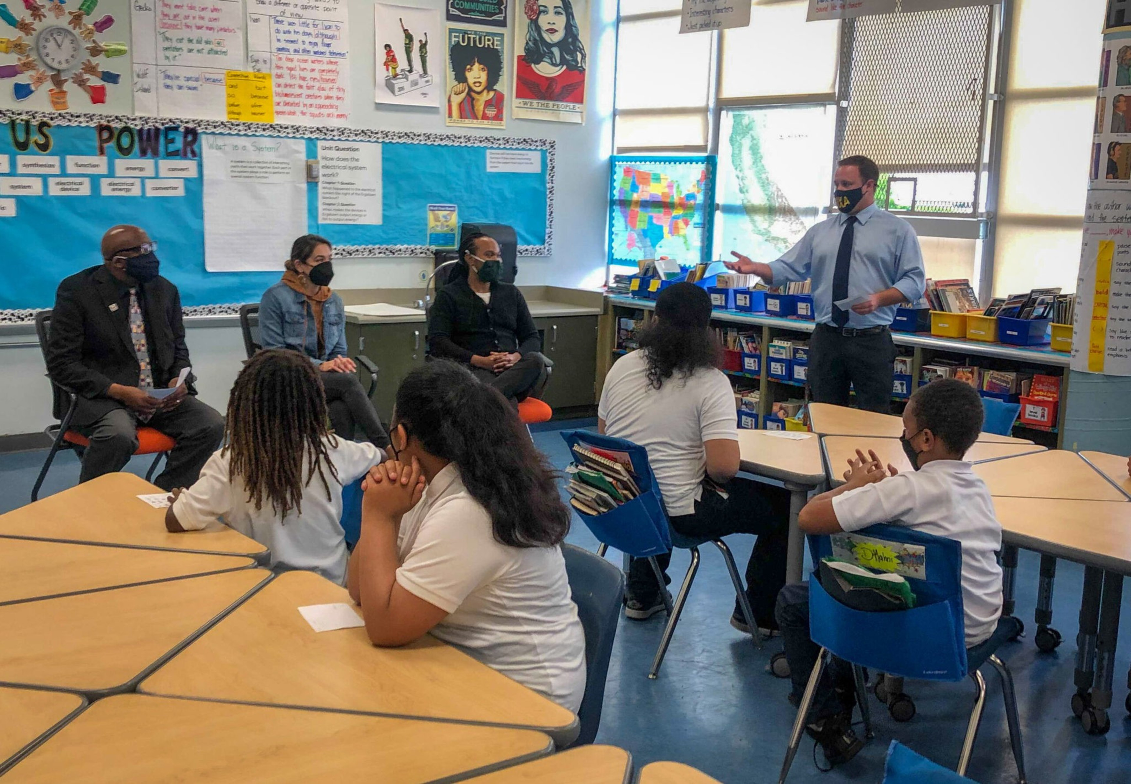 A teacher wearing a mask talks to students and three adults in a classroom decorated with posters and student work.