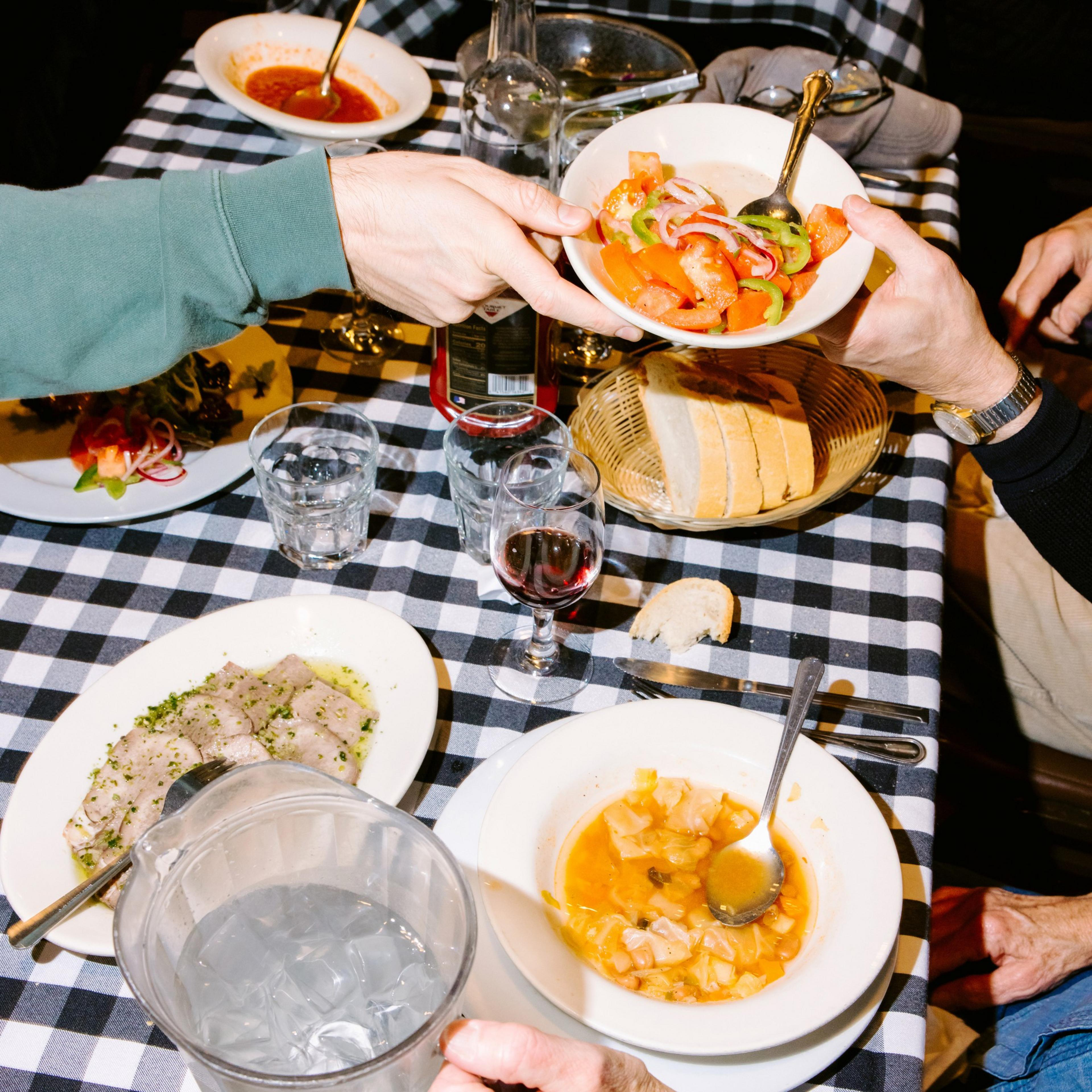 A checkered table holds plates of food, including a salad, soup, and sliced bread. Two people pass a bowl of salad, with glasses of water and red wine nearby.
