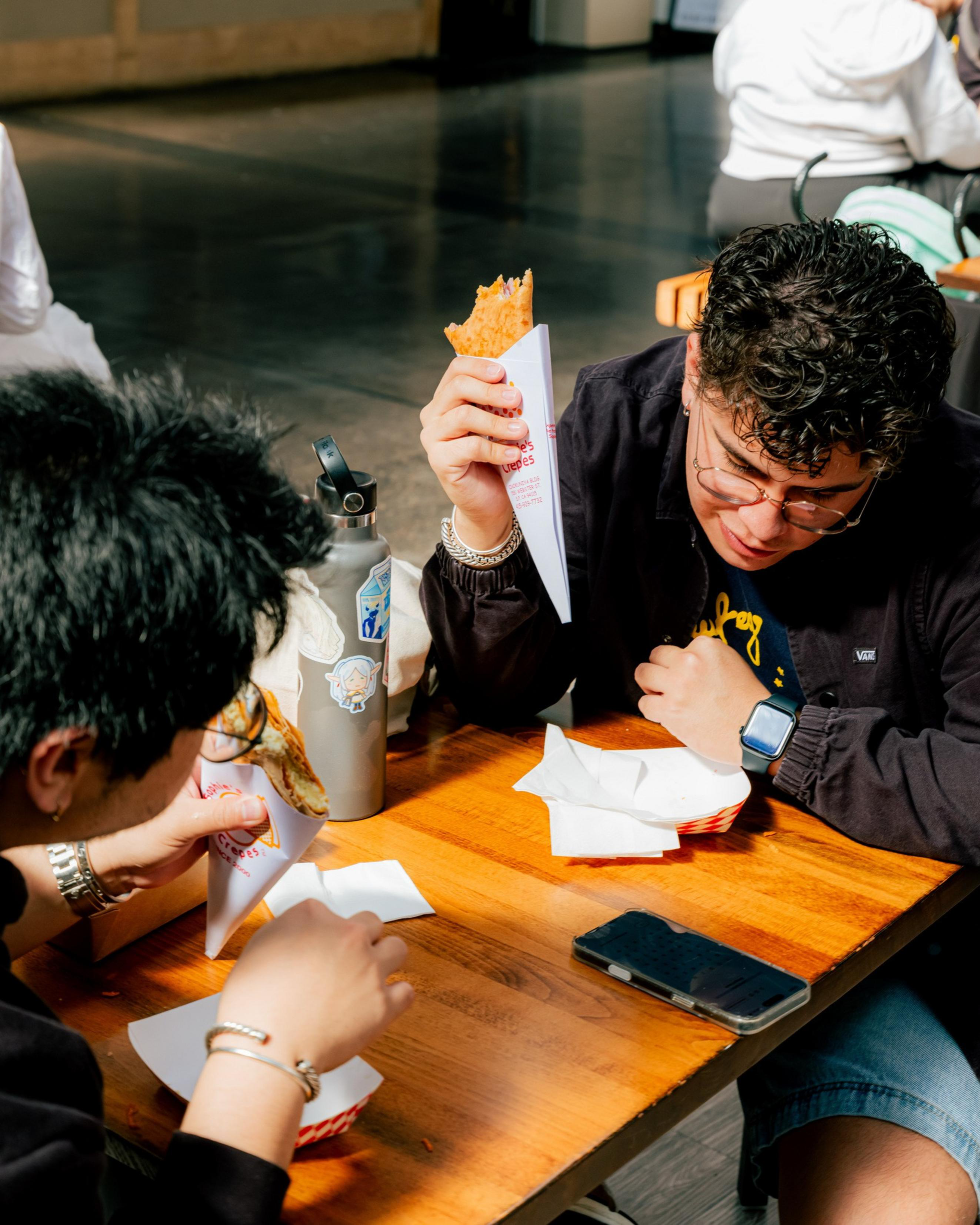 Two people sit at a wooden table eating food wrapped in paper cones, with a water bottle and a smartphone also on the table.
