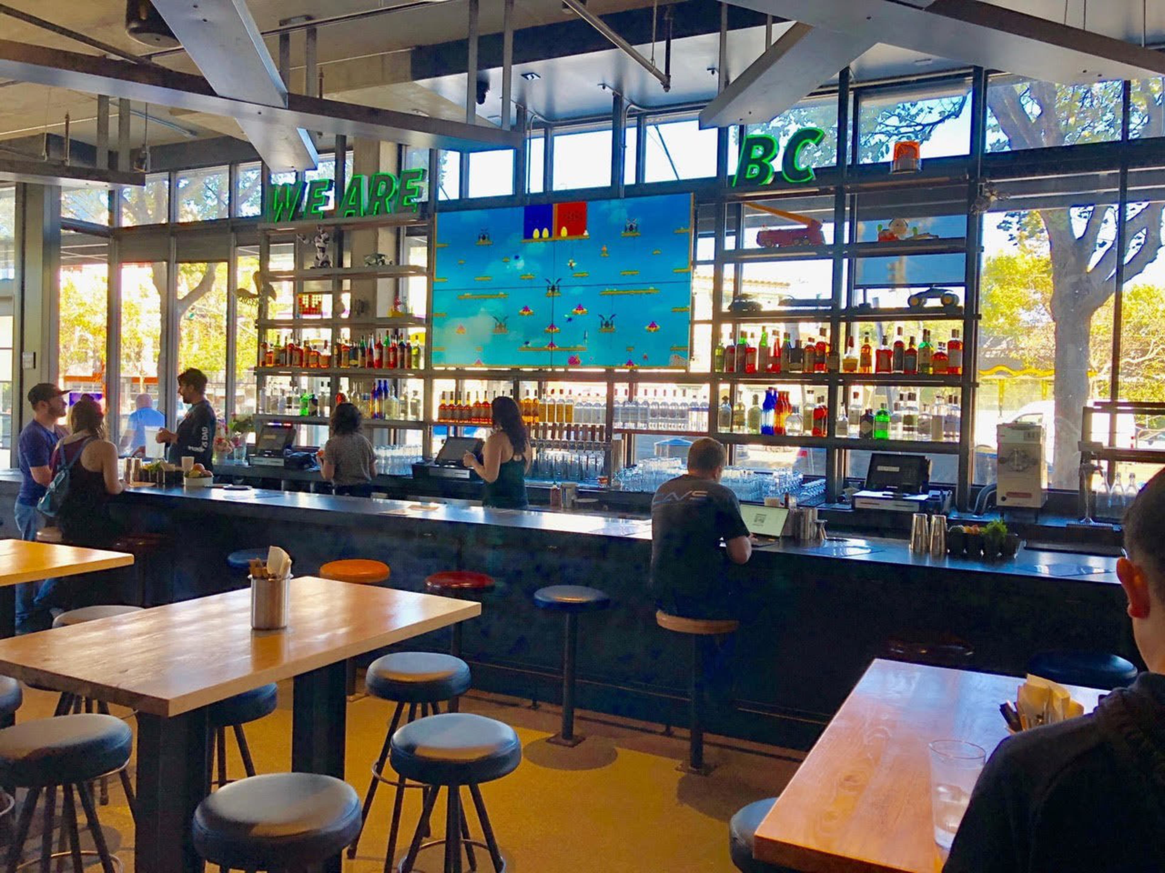 Customers sit at a bar on colored stools with tables in the foreground and a sign reading &quot;We Are BC&quot; in the background.