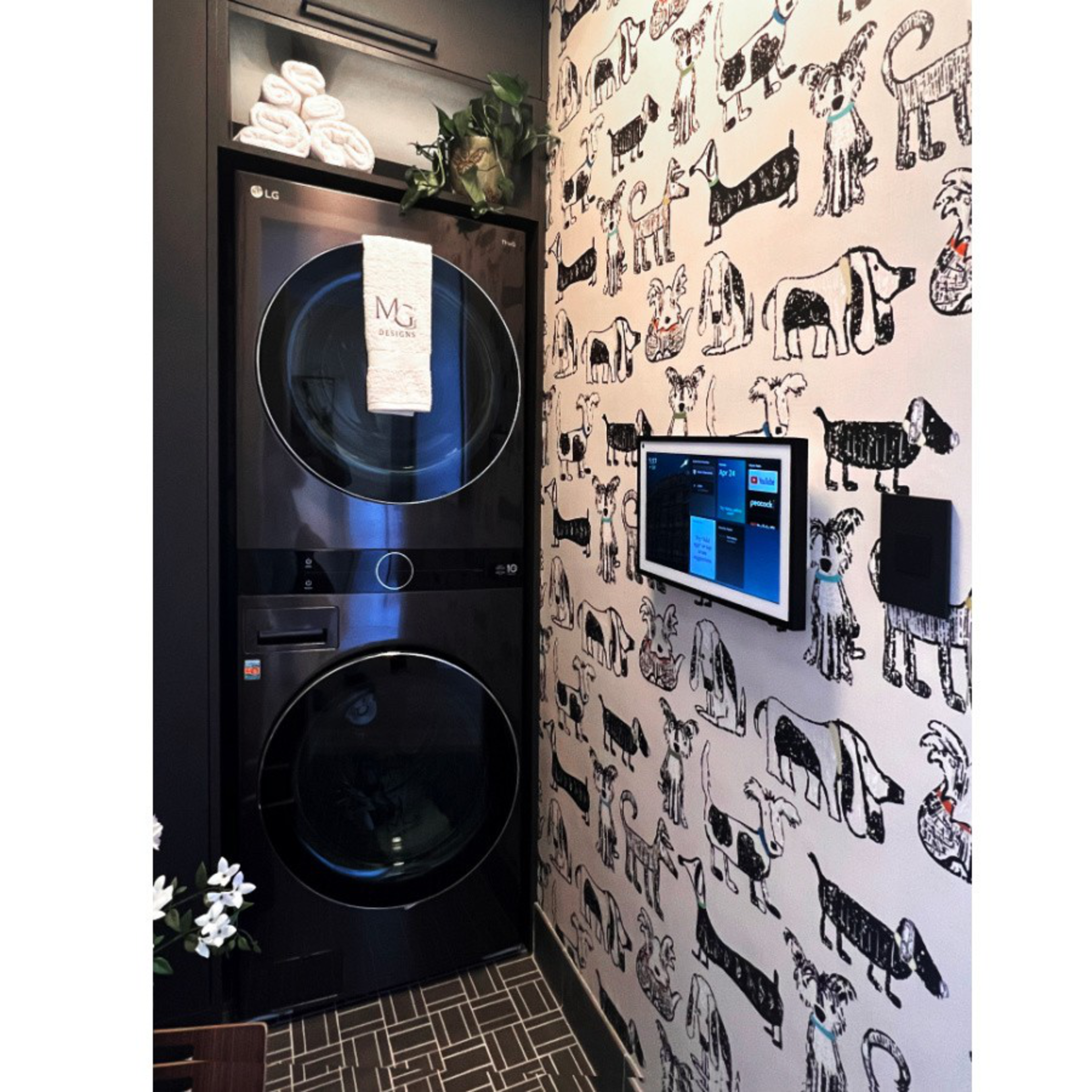 A stacked black washer and dryer sit beside a wall covered in playful black-and-white dog illustrations, with a small screen mounted on the wall.