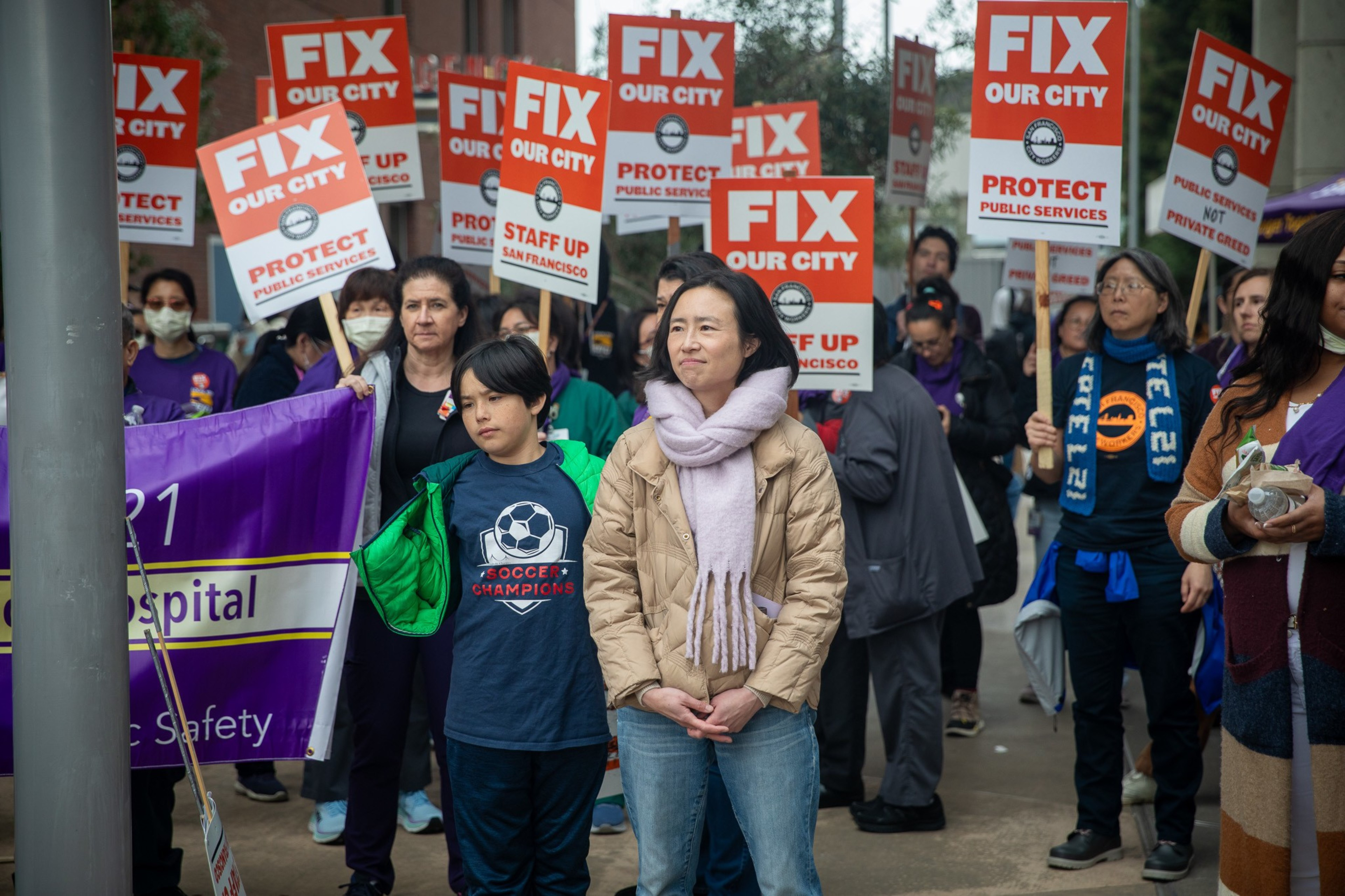 A crowd with &quot;FIX OUR CITY&quot; signs at a protest, a child in a soccer shirt looks forward.