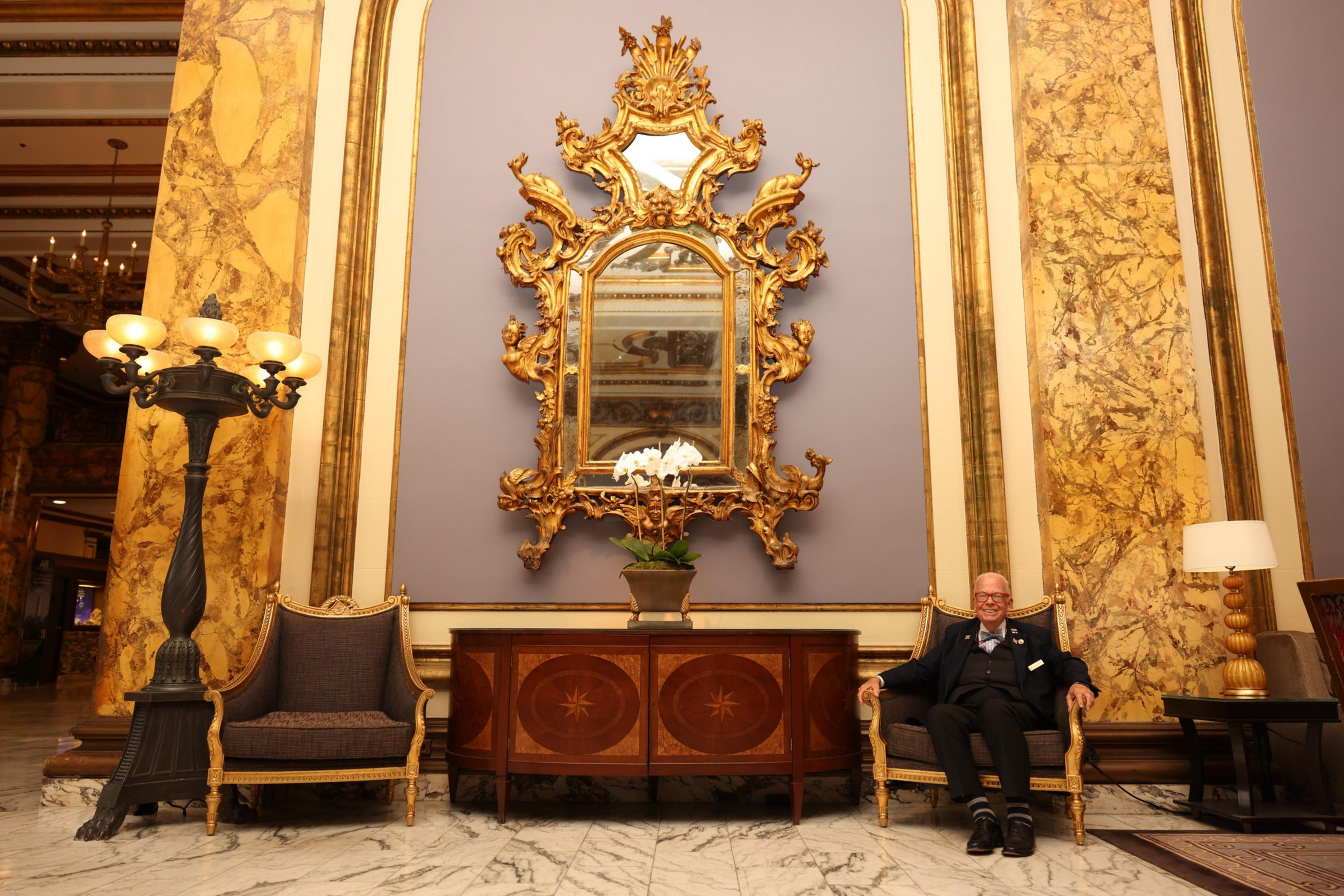 A man sits in the hotel by a wall with an ornate mirror.