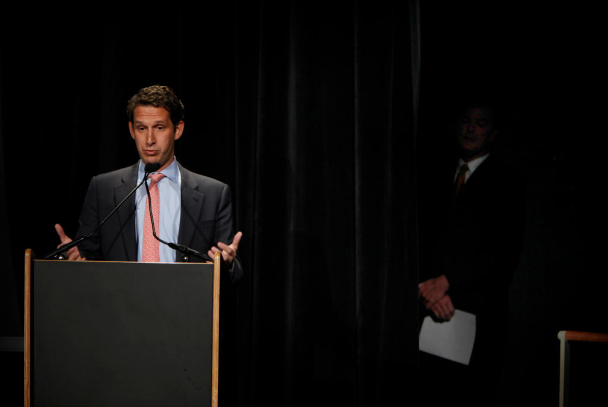 A man in a suit and pink tie speaks at a podium with microphones, while another man in a dark suit stands partially hidden behind a curtain holding papers.