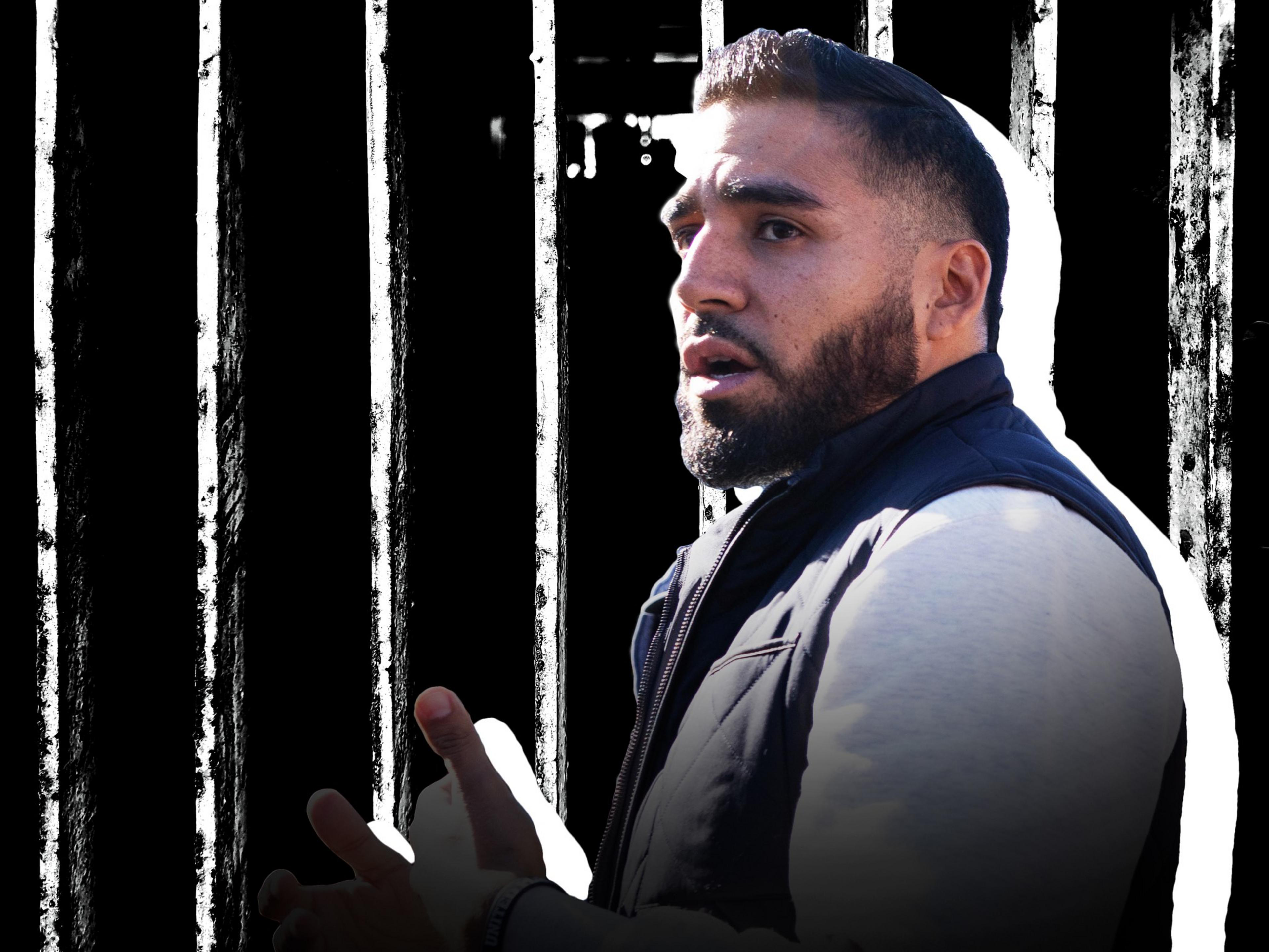 A man with a beard, wearing a dark jacket, is captured in a side profile against a background of vertical black and white stripes, suggesting prison bars.