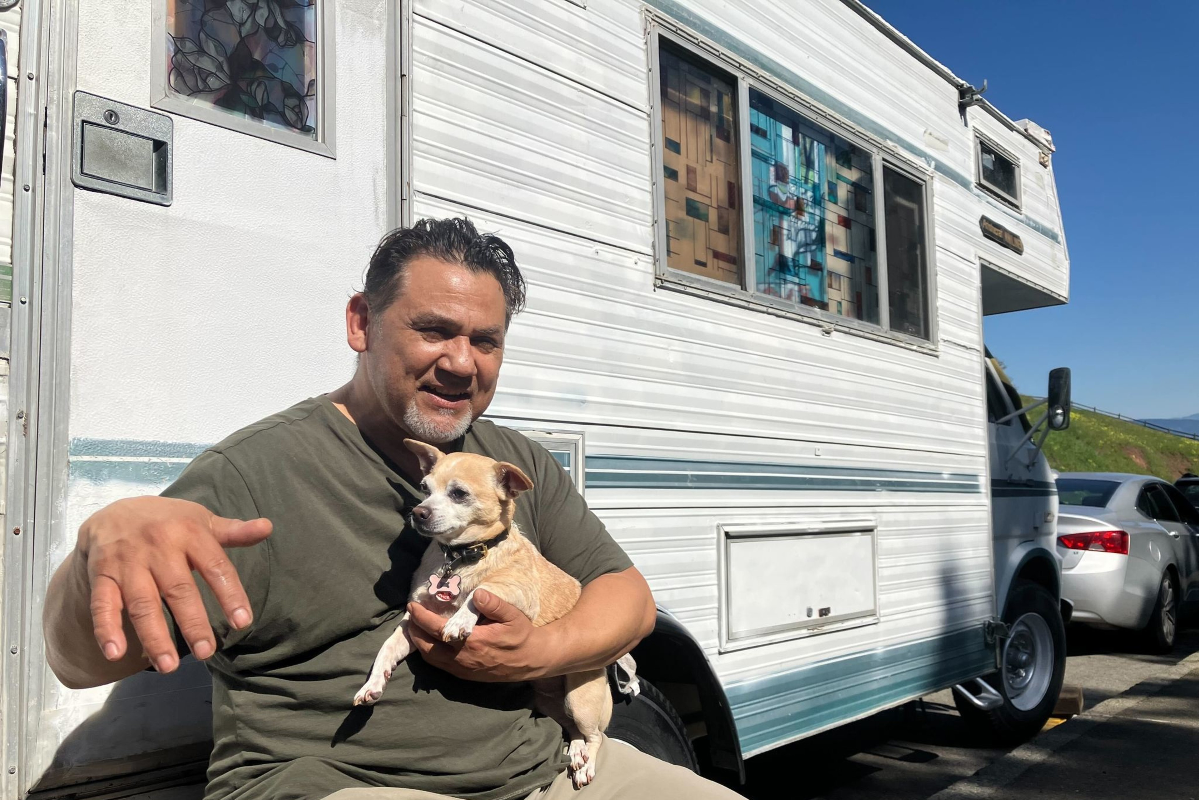 Man holds dog while sitting in front of white RV parked along street