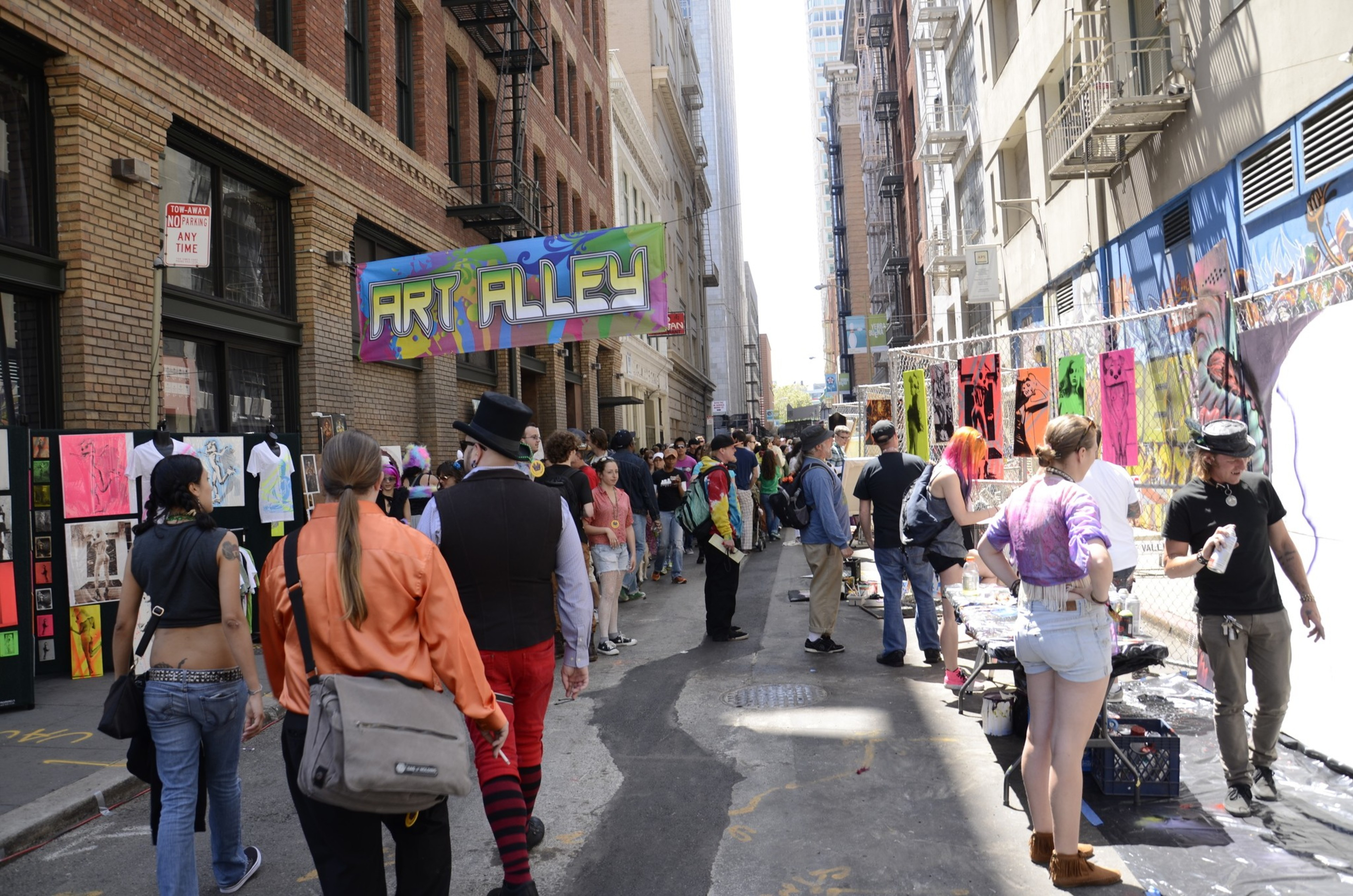 A bustling alley with colorful artwork on display. People walk and interact, some wearing colorful outfits. A vibrant &quot;Art Alley&quot; sign hangs overhead.