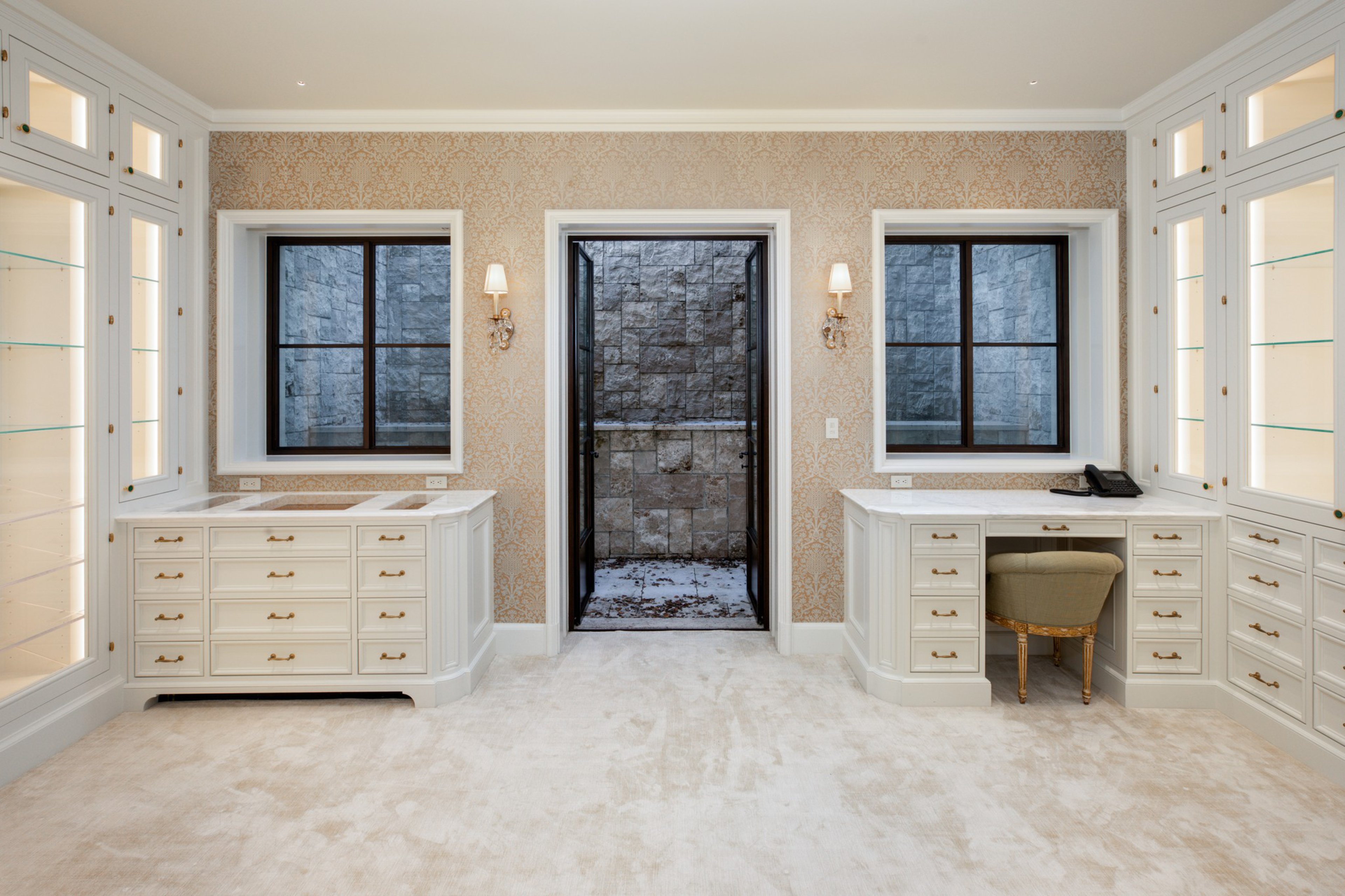 A room with cream cabinetry and drawers, two windows, a central open doorway to a stone outdoor area, gold-patterned wallpaper, beige carpet, and two wall sconces.