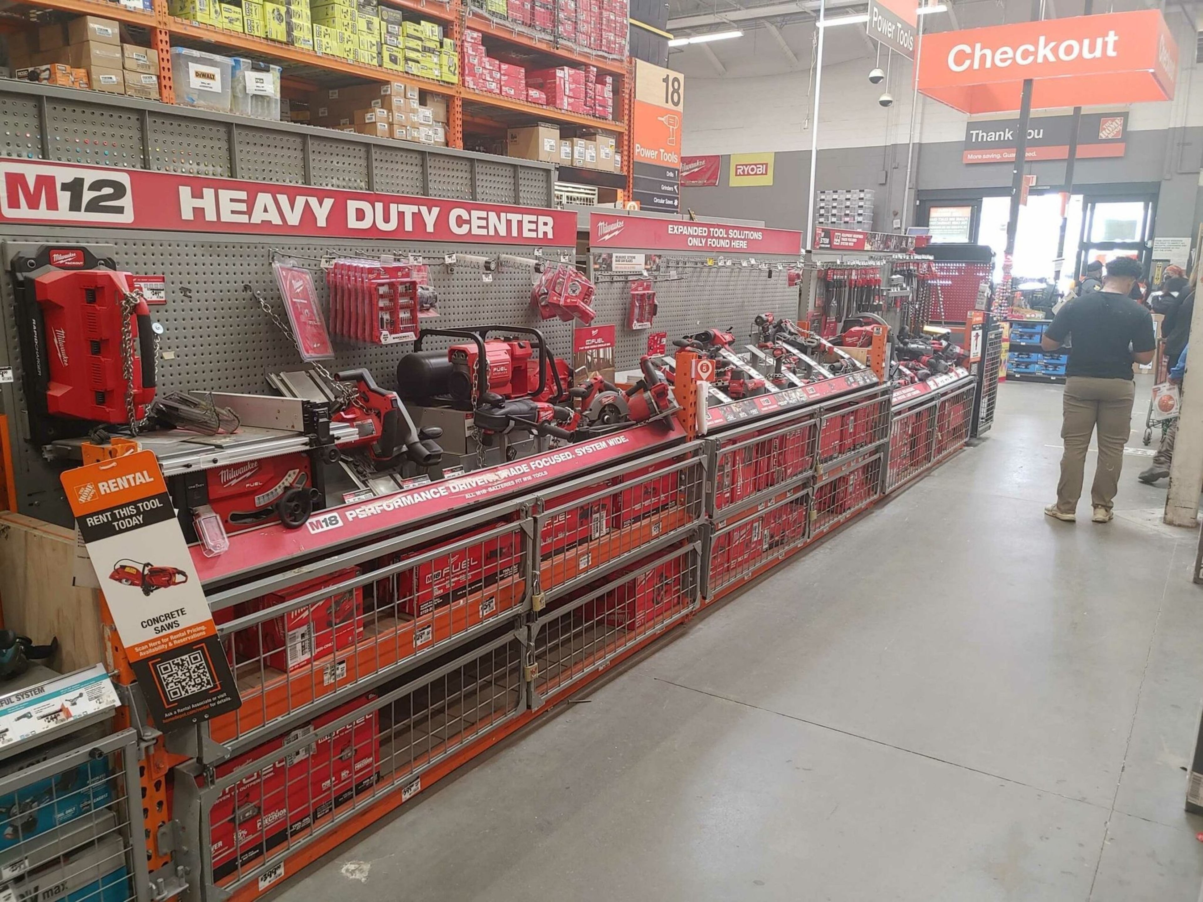 A hardware store aisle features a long display of Milwaukee power tools labeled "Heavy Duty Center," with a checkout area visible in the background.