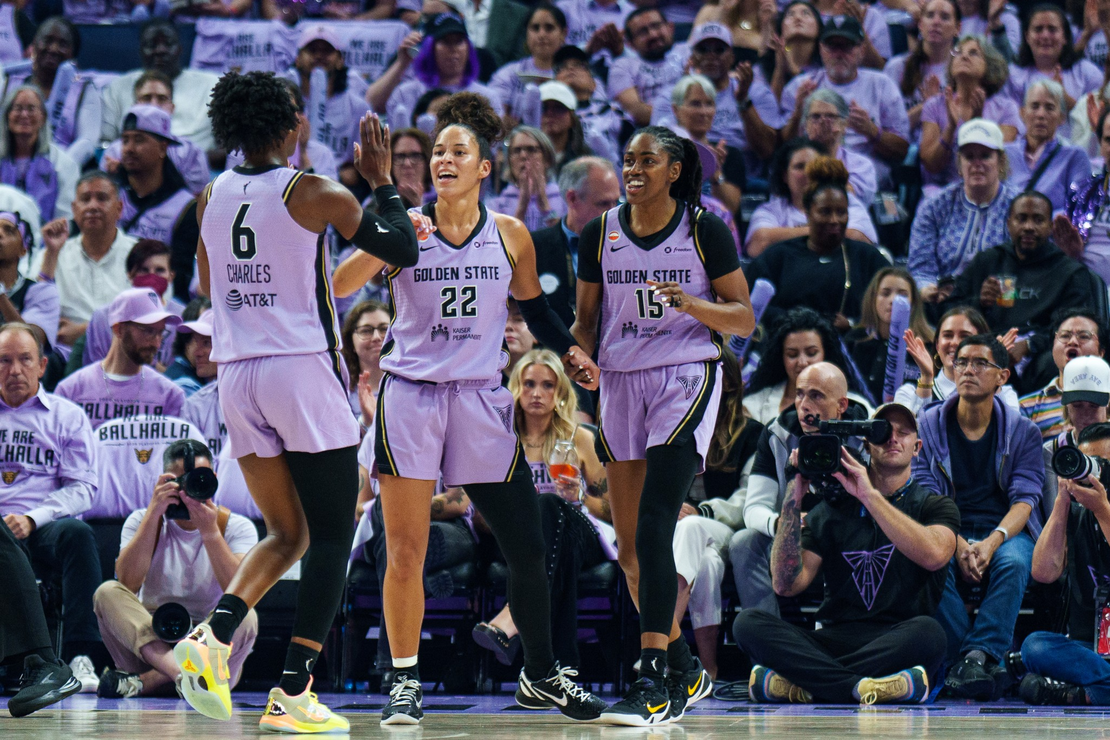 Three Golden State basketball players in purple uniforms celebrate on court while a seated crowd and photographers watch closely.