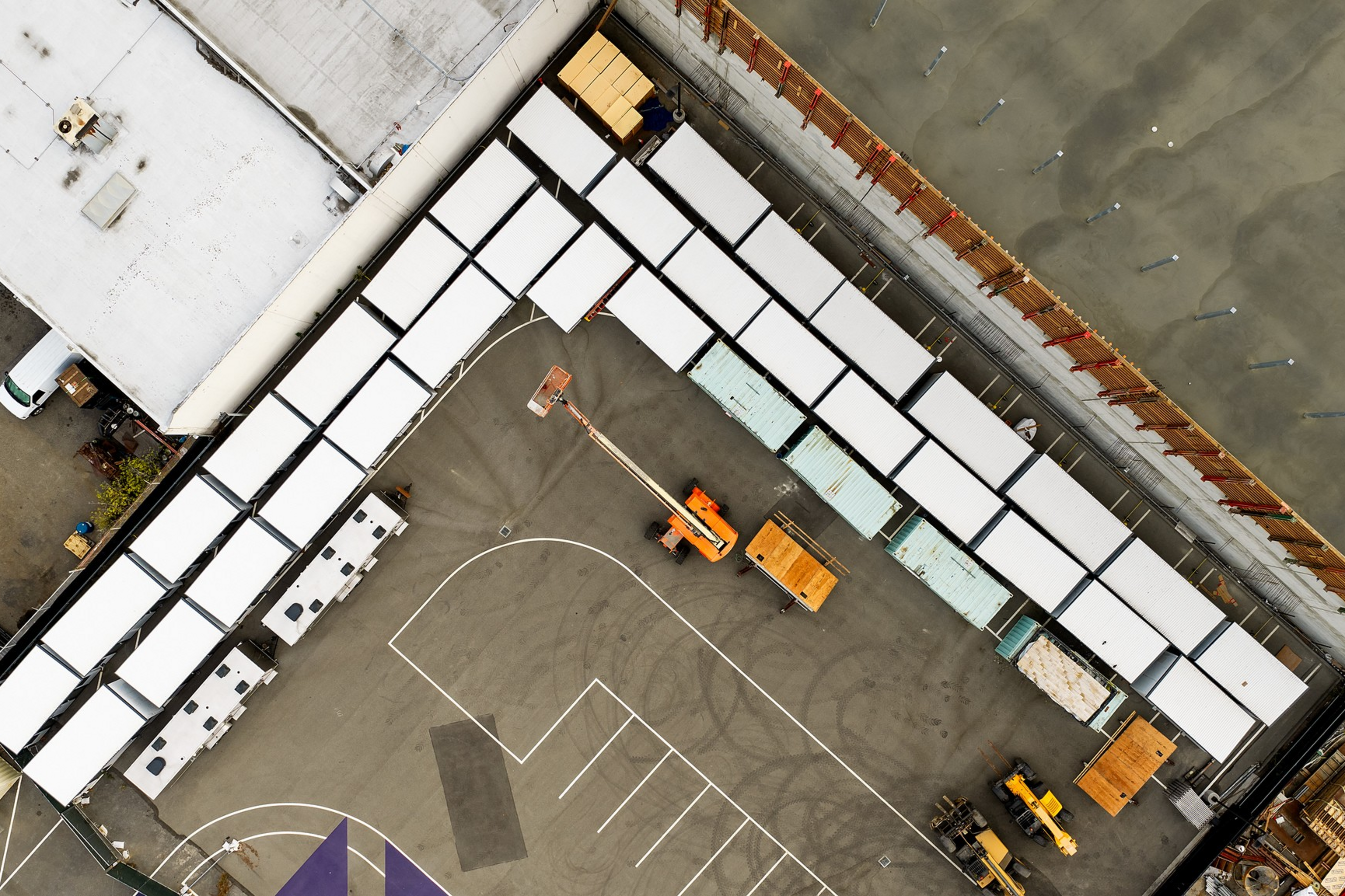 Aerial view of worksite with white trailers lined up in an L-shape, two orange construction vehicles, a forklift, and parking spaces on asphalt.