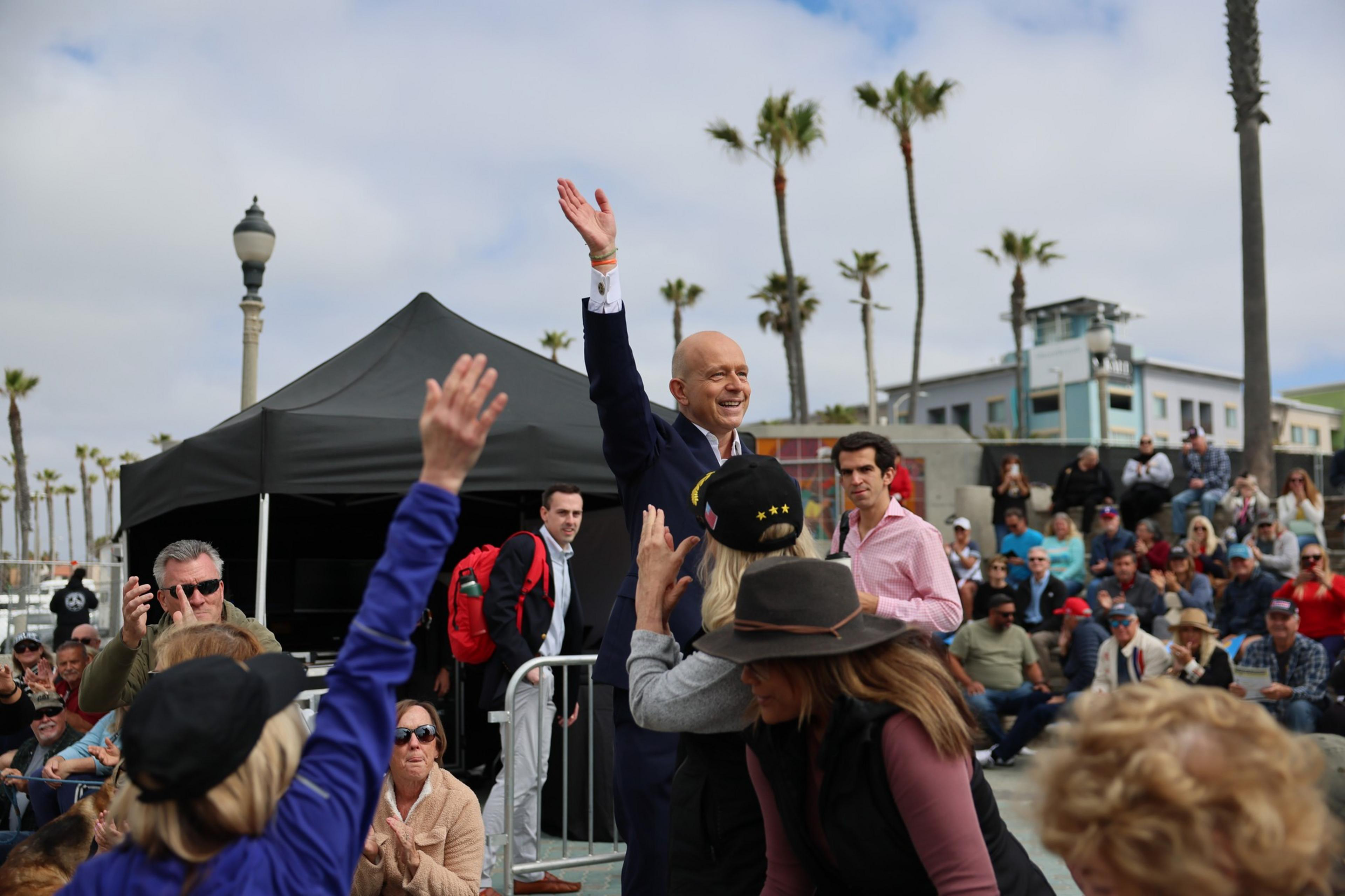 A man waves to a crowd at an outdoor event by the beach. People surround him, clapping and raising their hands. Palm trees and a tent are in the background.