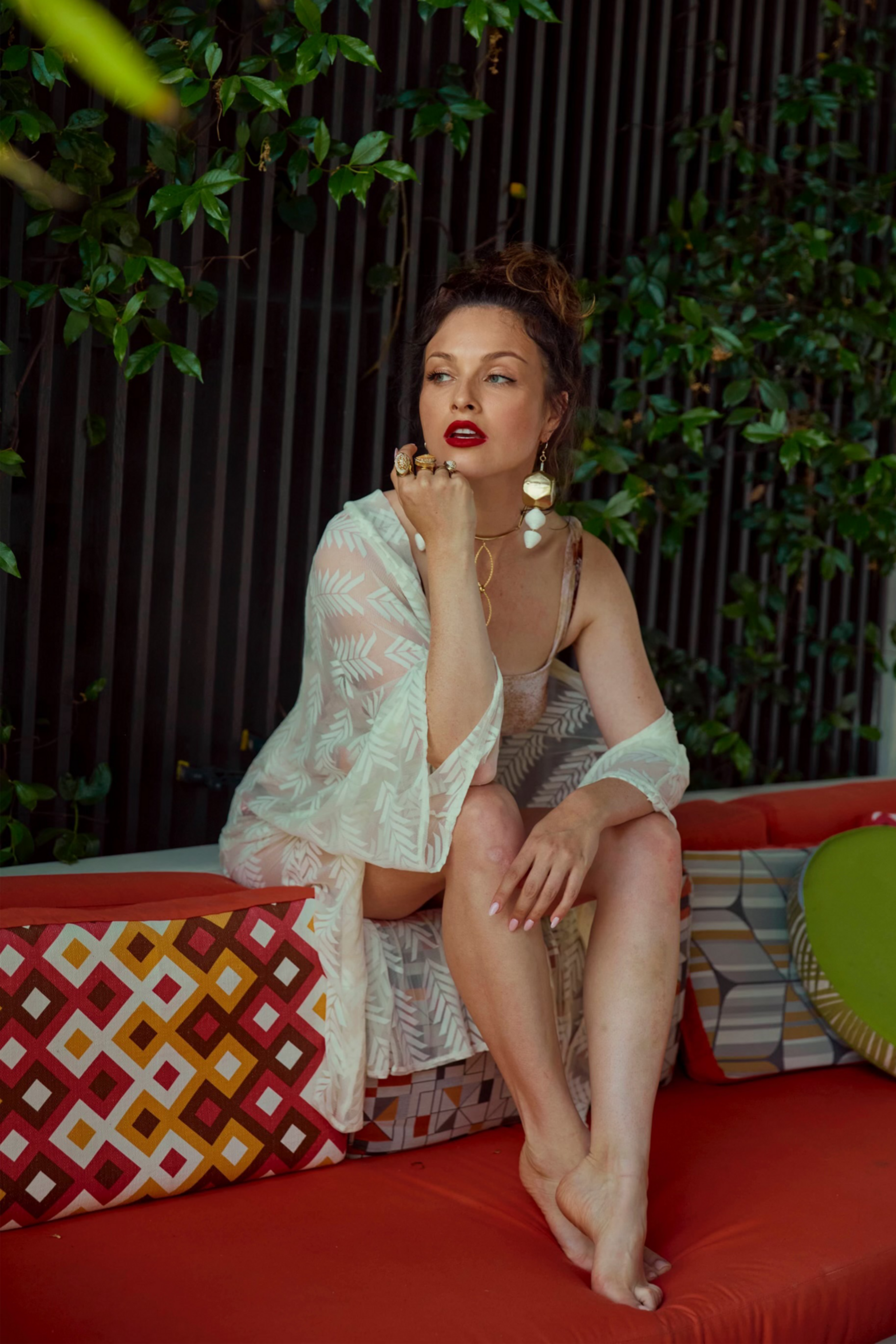 A woman sits on a patterned couch with a leafy background. She wears a sheer wrap, chunky jewelry, red lipstick, and has her hair up, looking thoughtfully aside.