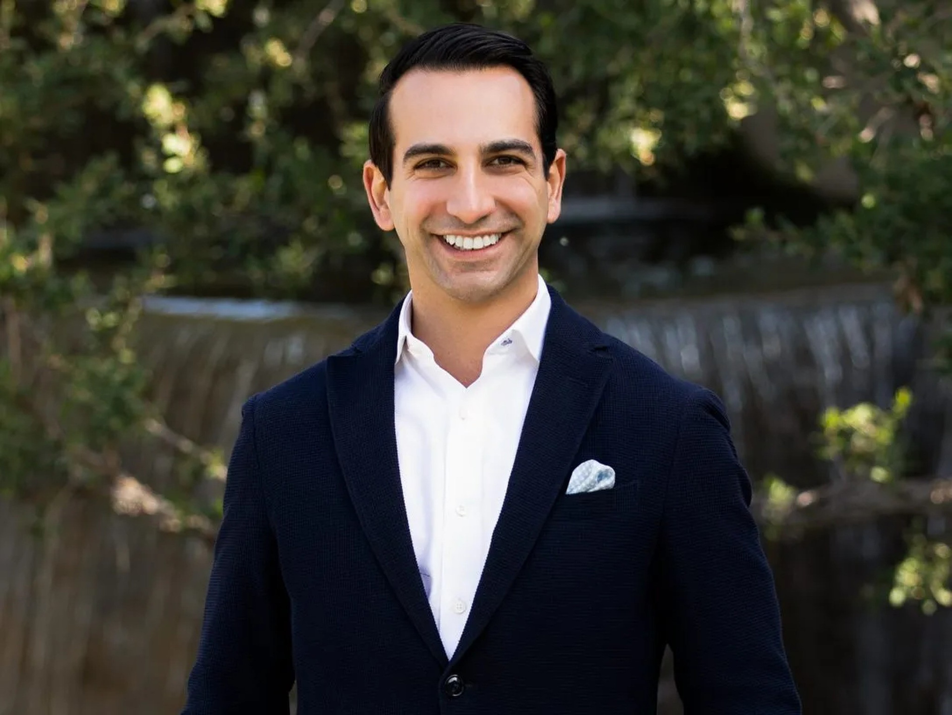 A man in a dark suit and white shirt smiles outdoors, with a blurred green foliage and waterfall background.