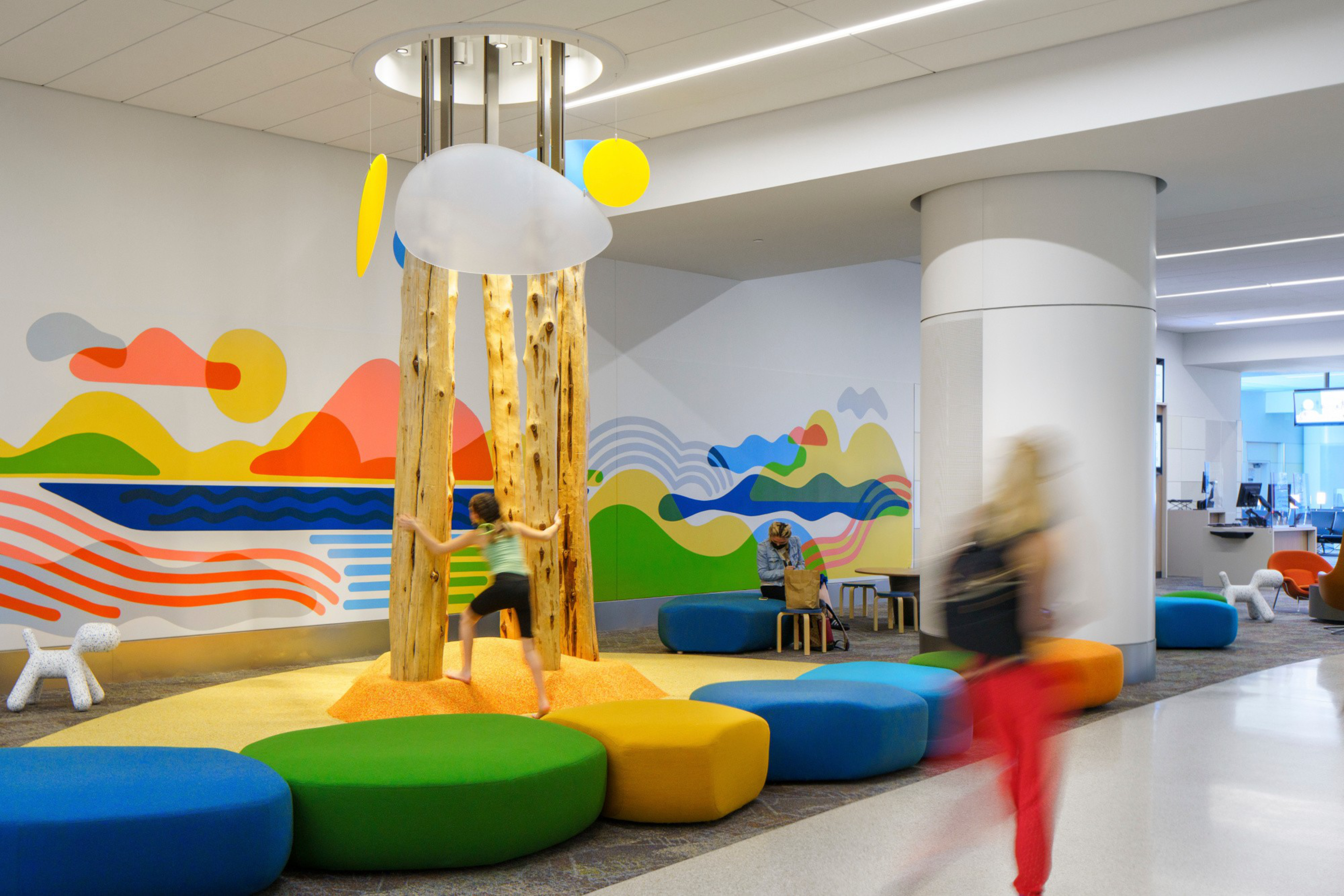 A colorful indoor play area has a wooden tree structure with hanging clouds and lights, surrounded by vibrant cushioned seating and a mural on the wall.