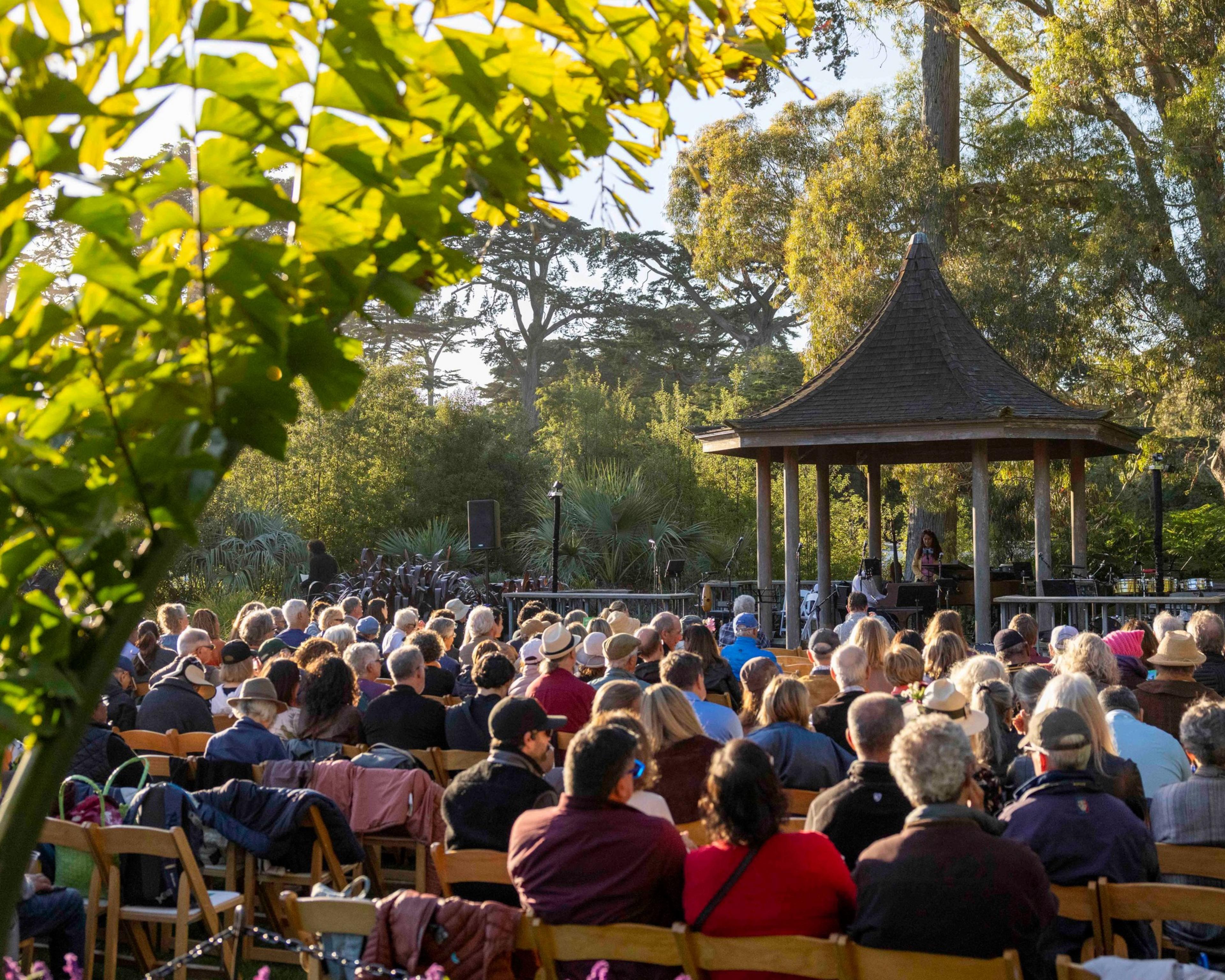 People are seated outdoors, facing a small wooden gazebo with a person speaking. Greenery and large trees surround the area, creating a peaceful setting.