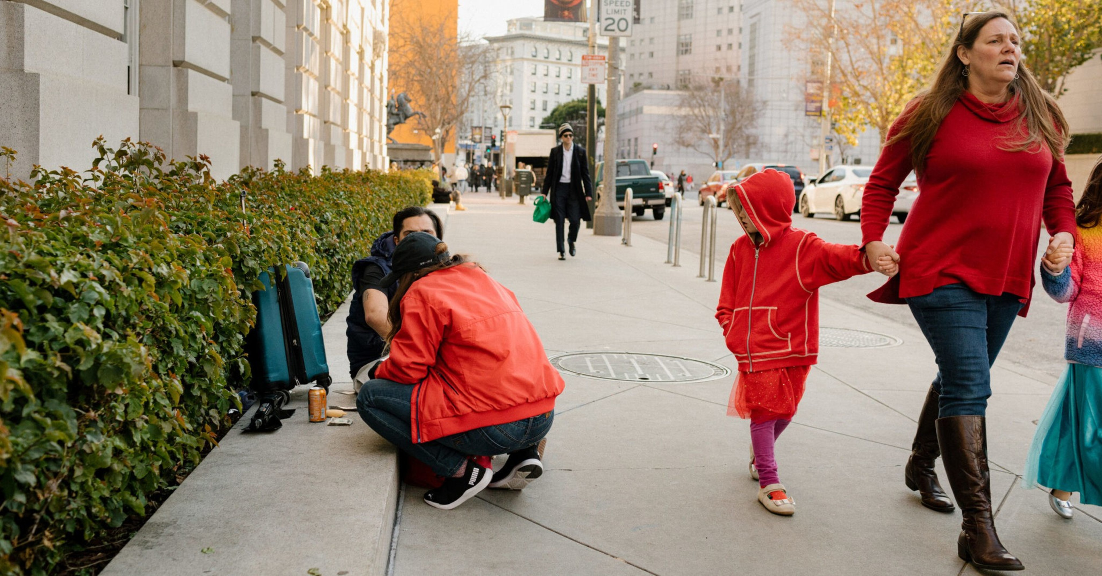Two people sit on a curb near green bushes, one wearing a red jacket, while a woman in a red sweater holds hands with two children walking on a city sidewalk.