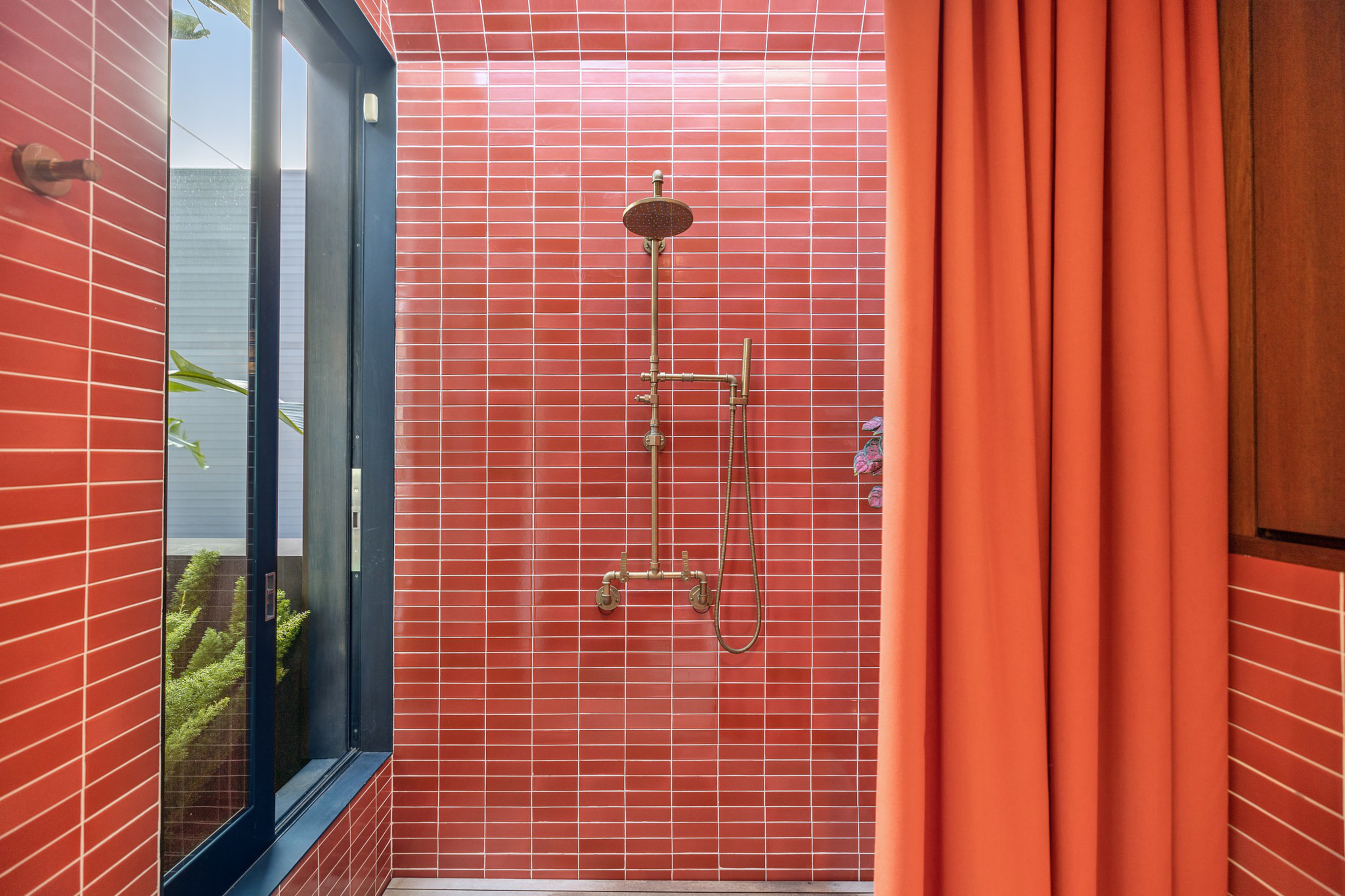 A shower with brass fixtures is set against red rectangular tiles, next to a large window with plants outside and an orange shower curtain.