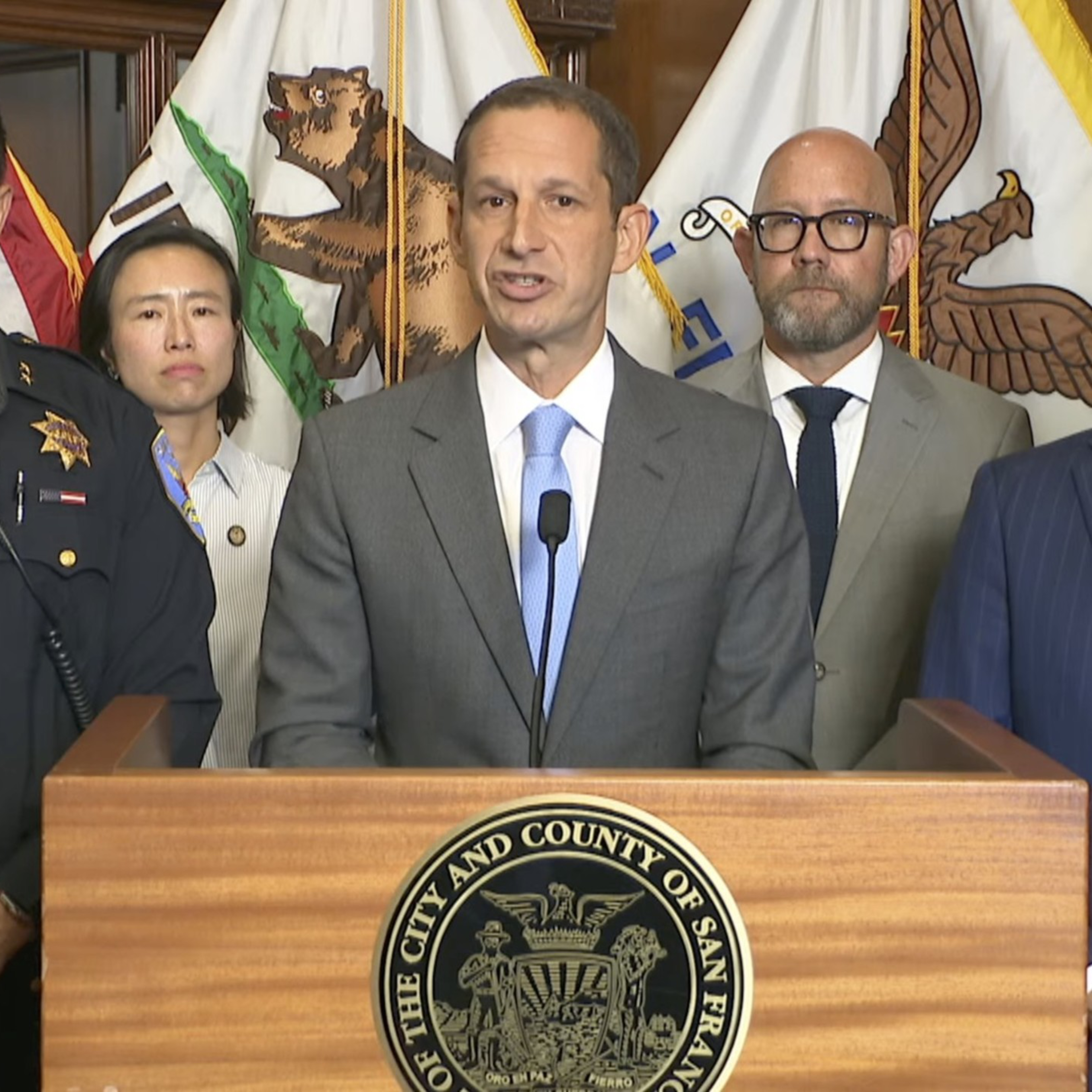 A man in a gray suit speaks at a podium with San Francisco seals, flanked by officials and police officers in front of American and California flags.