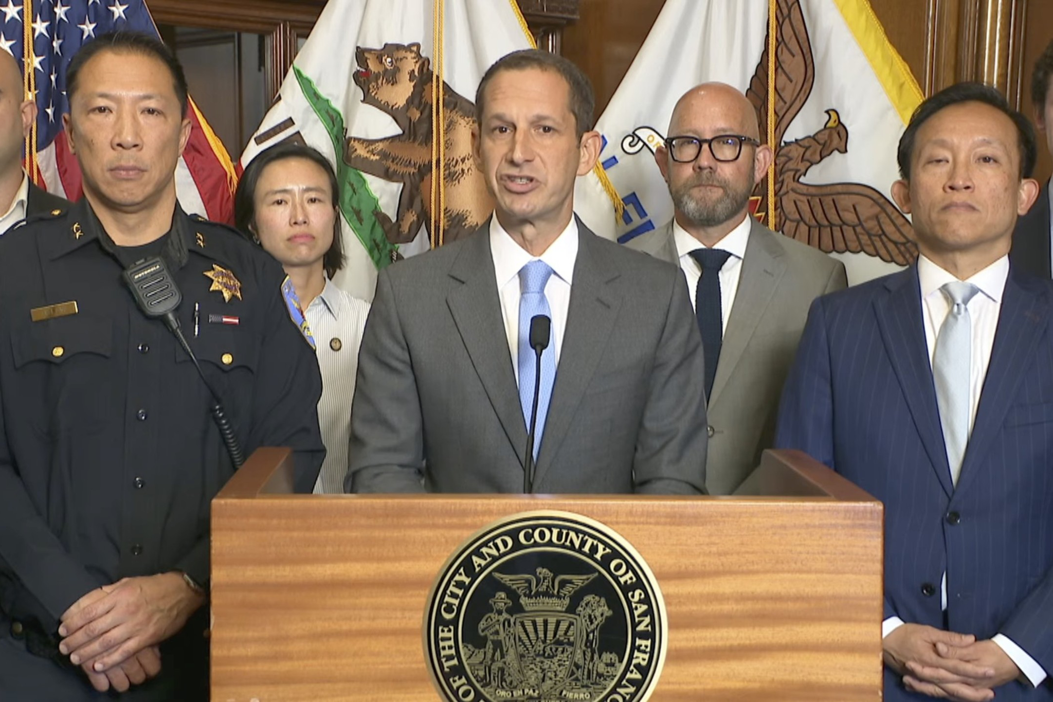 A man in a gray suit speaks at a podium with San Francisco seals, flanked by officials and police officers in front of American and California flags.