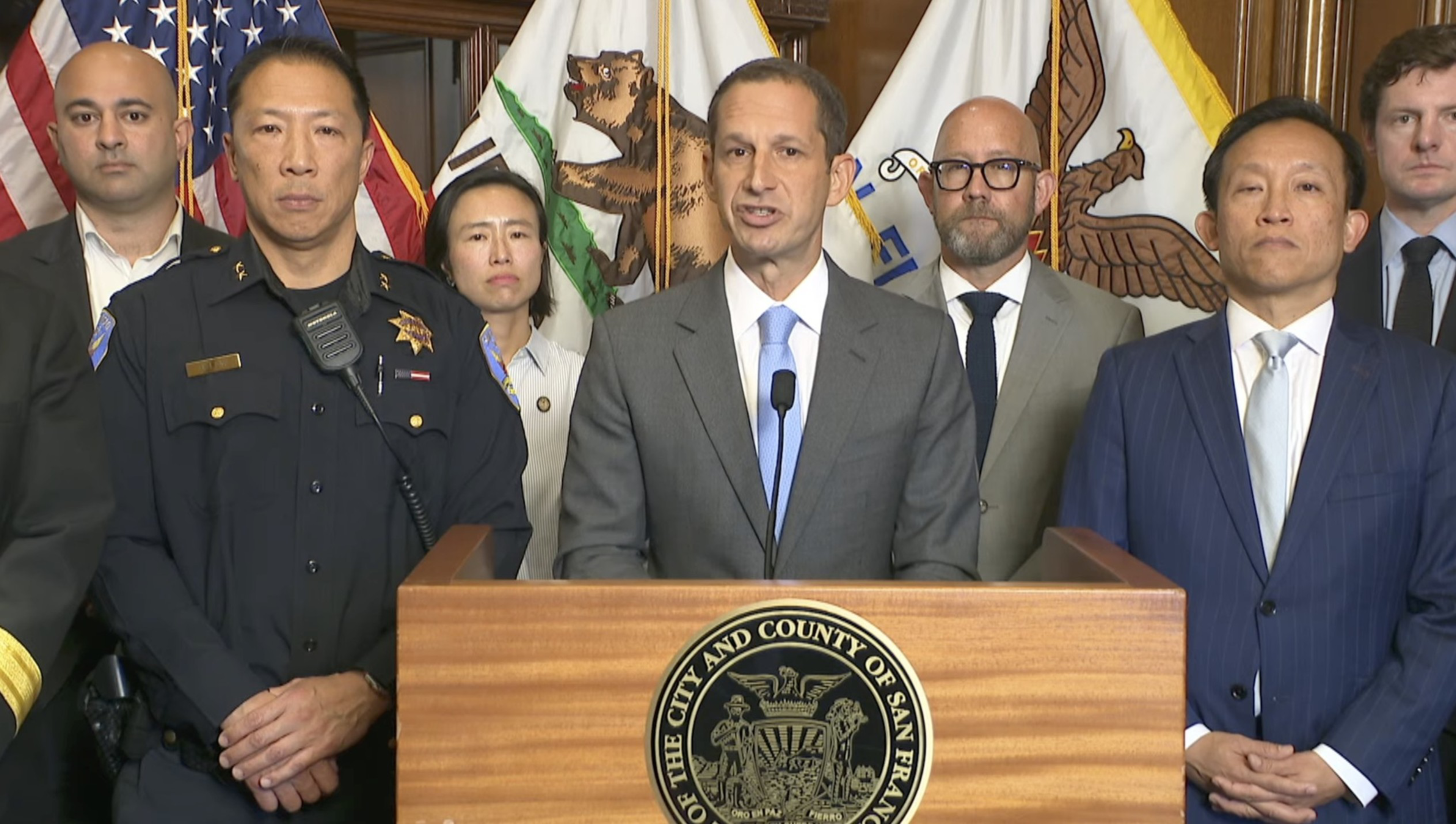 A man in a gray suit speaks at a podium with San Francisco seals, flanked by officials and police officers in front of American and California flags.
