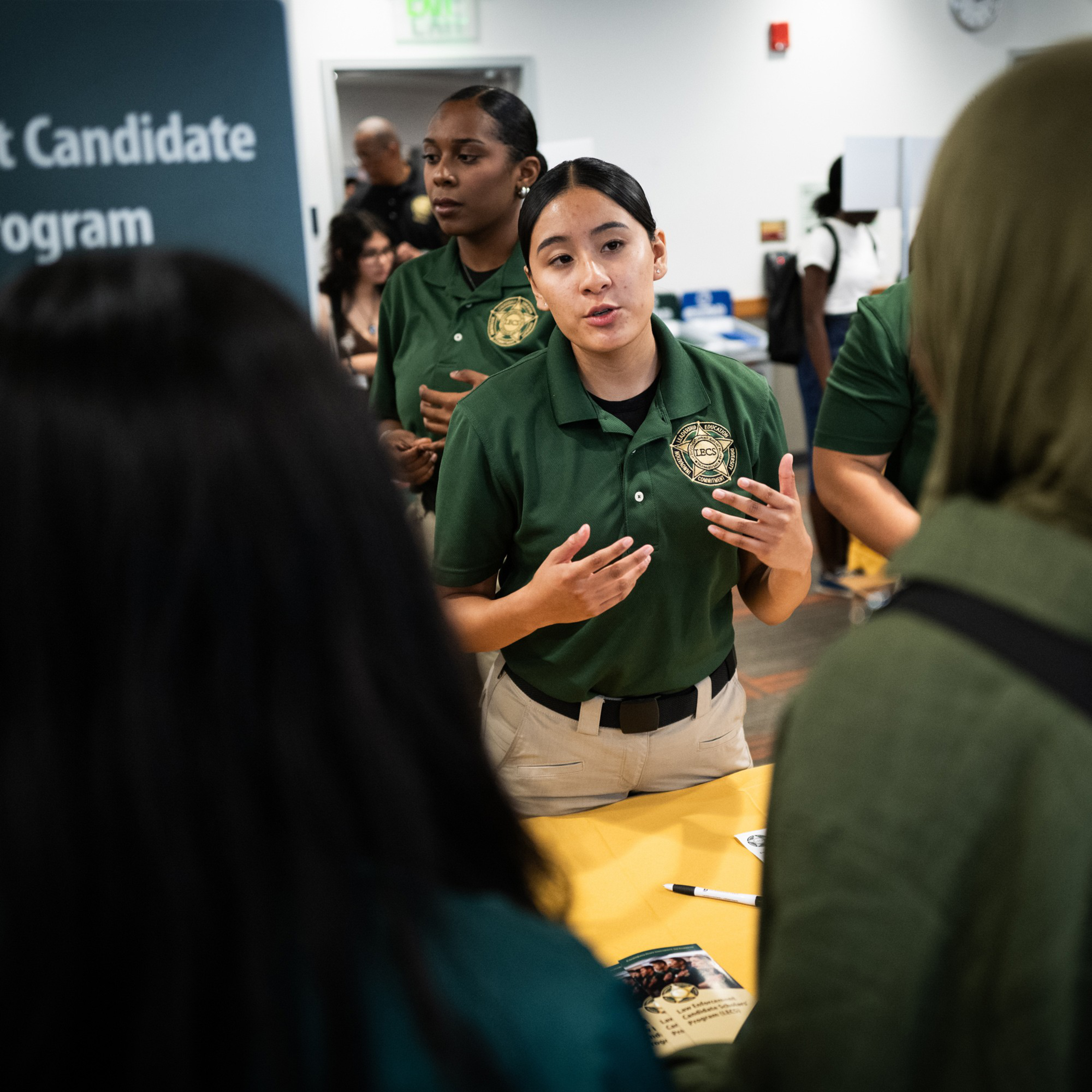 A uniformed woman speaks to two people at a table. Other uniformed individuals and attendees are visible in the background. A sign reads &quot;Candidate Program.&quot;