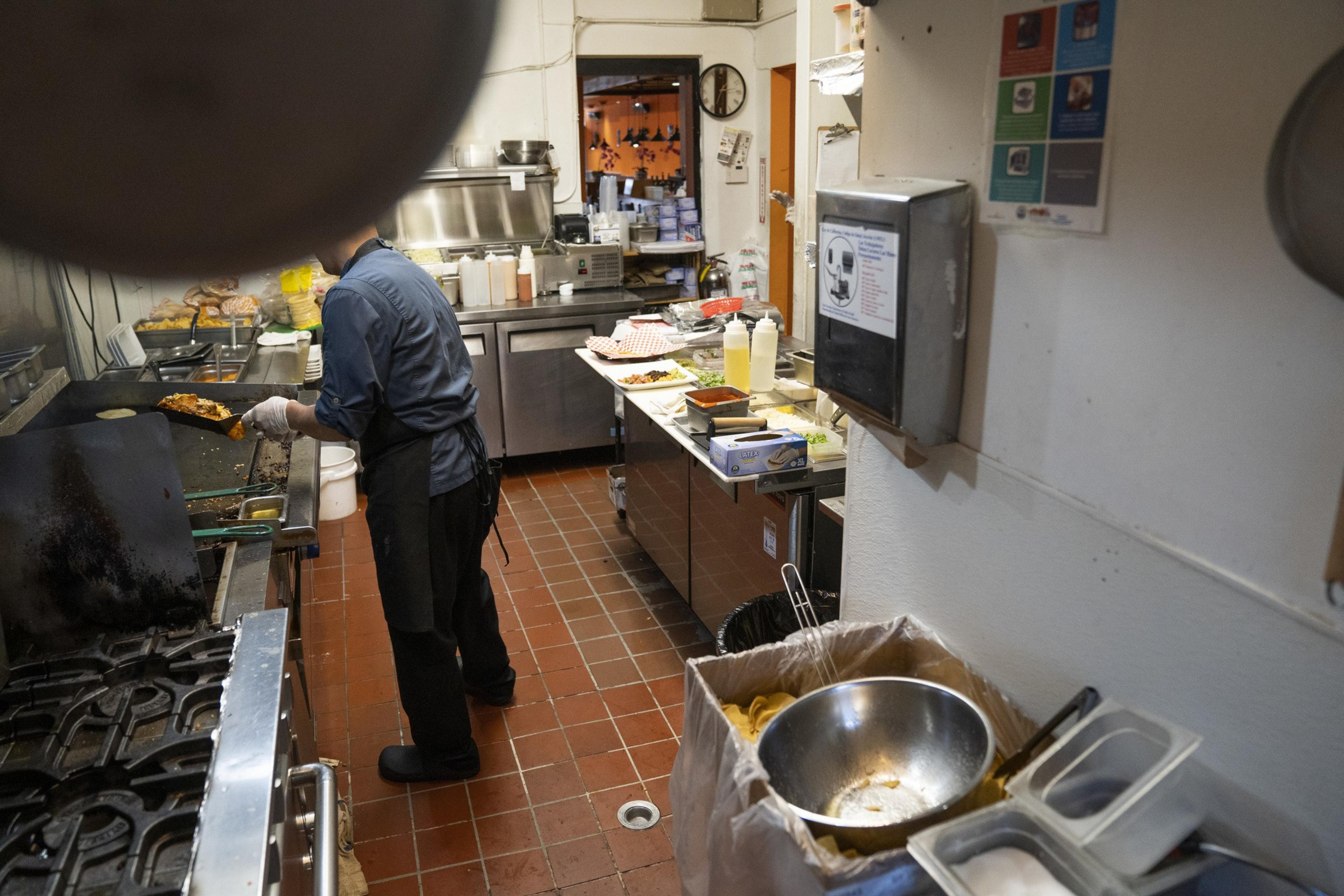 A cook in a commercial kitchen prepares food, standing by a stove. The space is filled with utensils, containers, and food ingredients on a countertop.