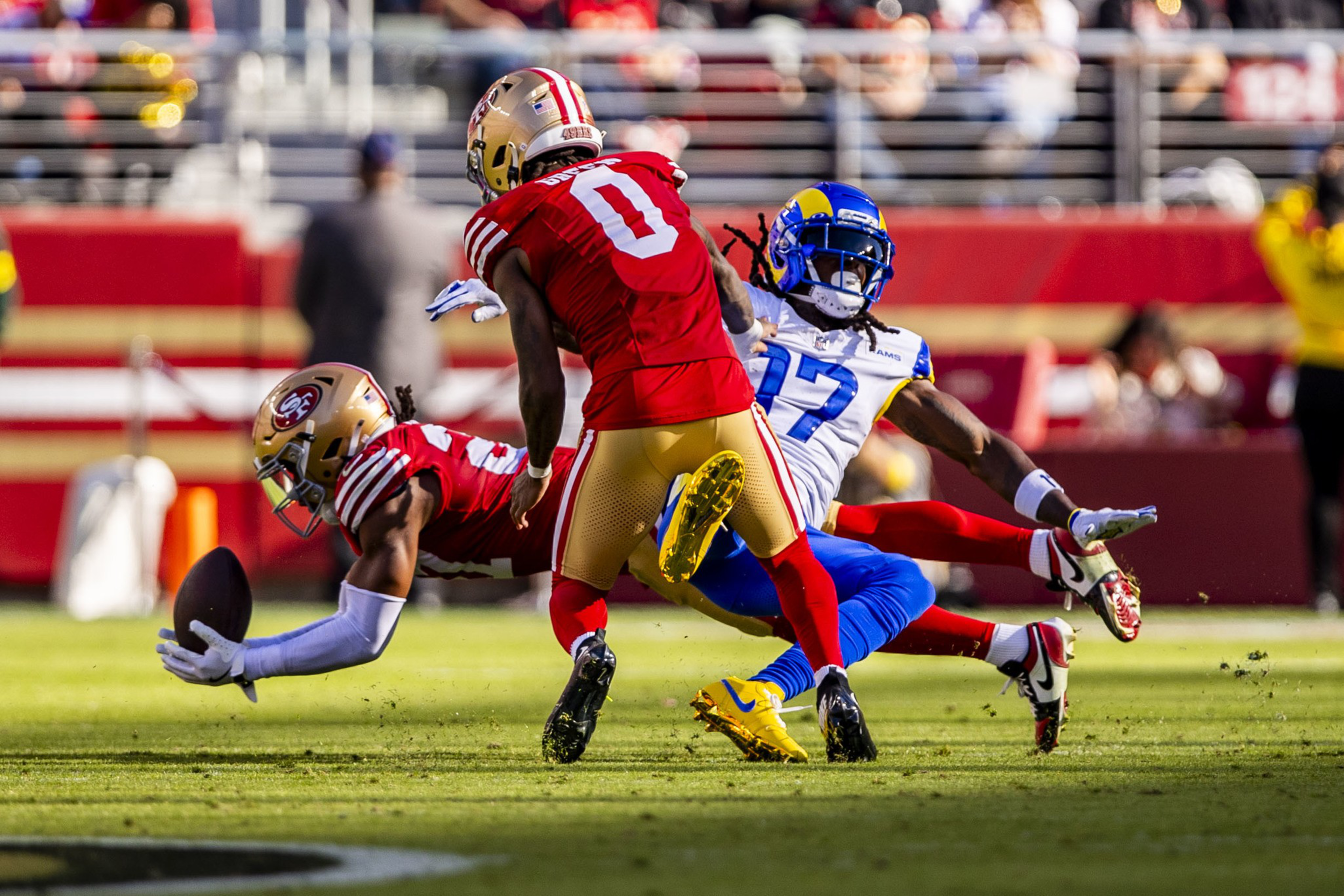 A football player in a red uniform dives to catch the ball near the ground while another red-uniformed player blocks a blue-uniformed opponent falling backward.