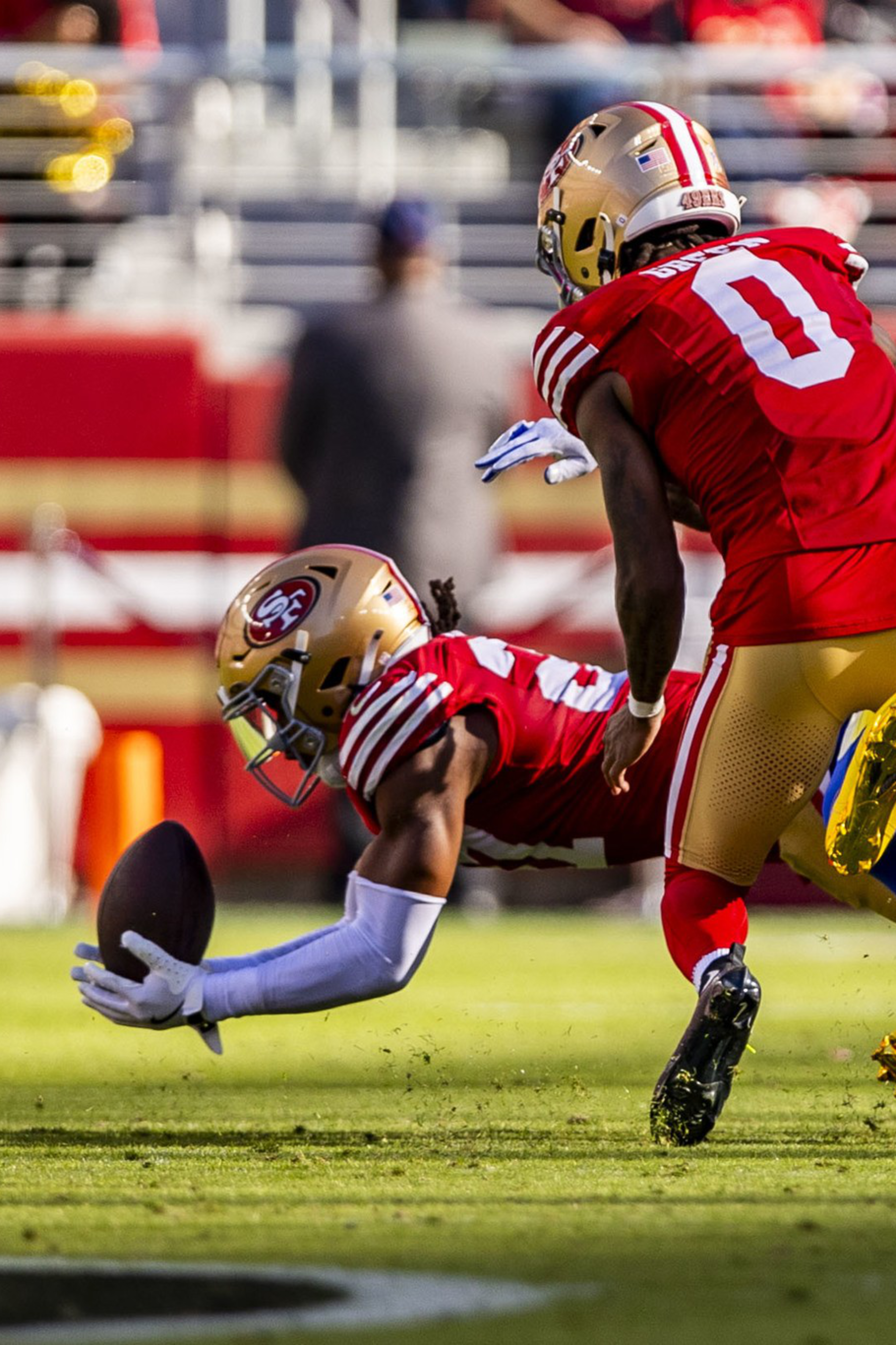 A football player in a red uniform dives to catch the ball near the ground while another red-uniformed player blocks a blue-uniformed opponent falling backward.