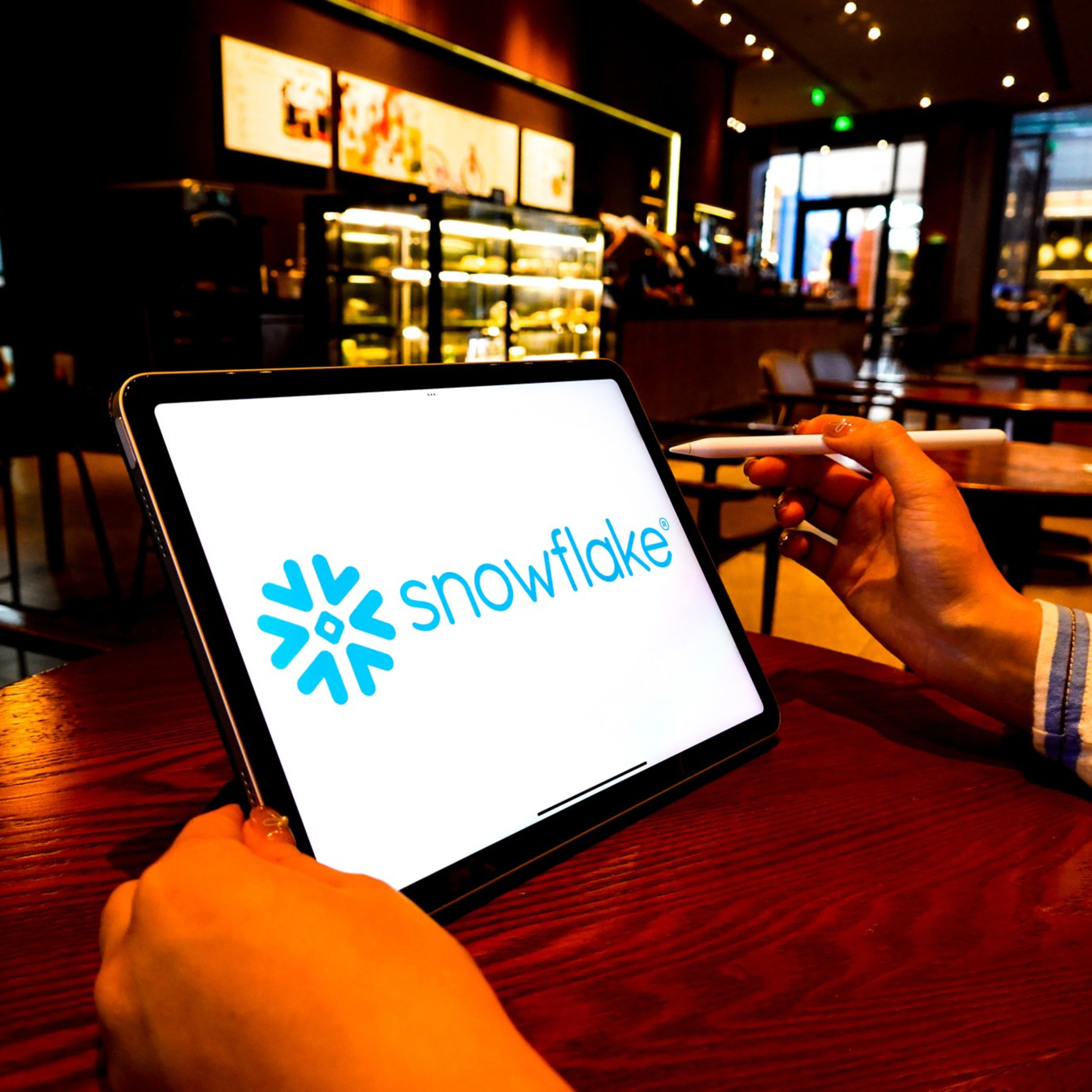 A person holds a tablet displaying the Snowflake logo, seated at a wooden table in a cozy, well-lit cafe with a modern interior.