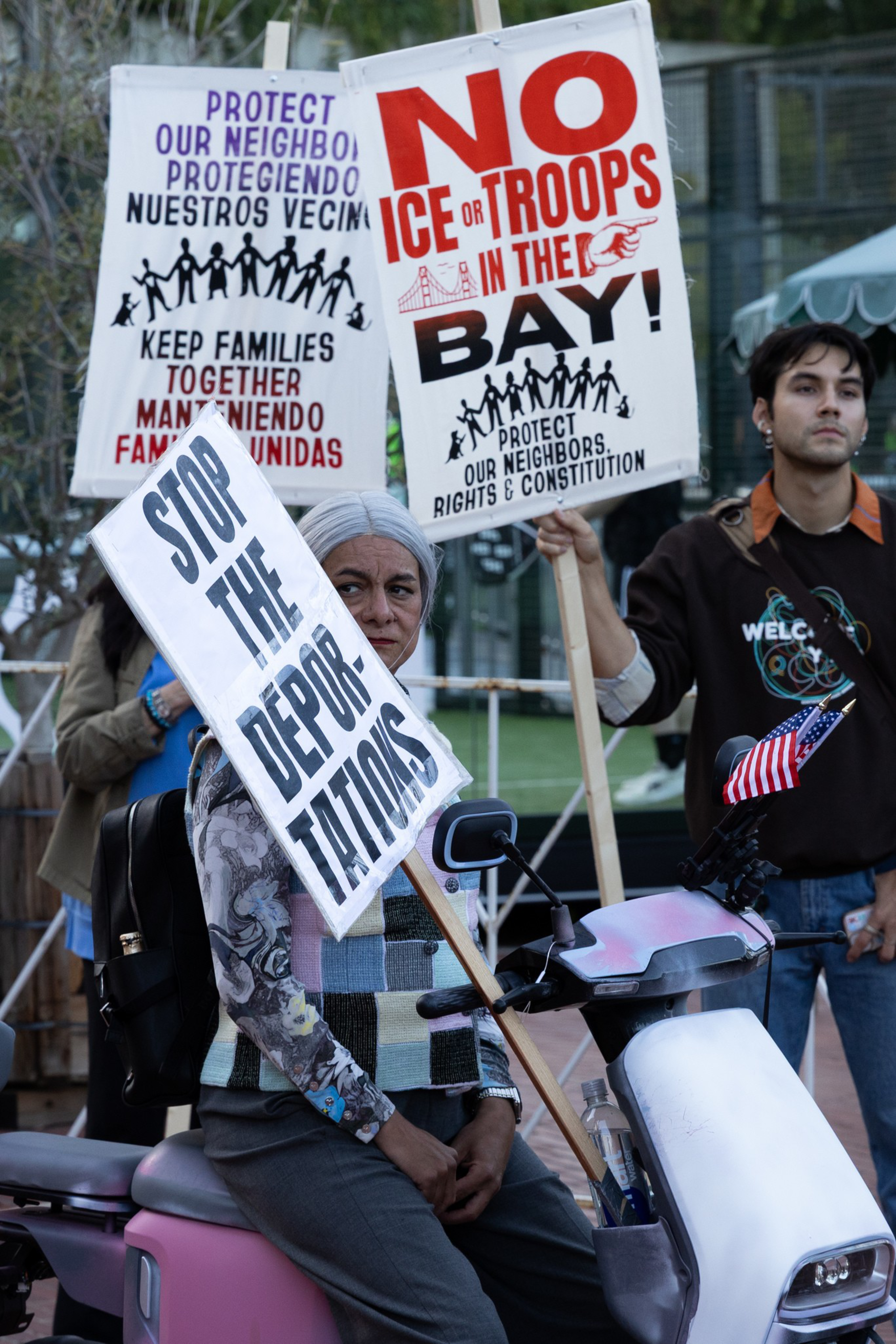 People hold signs protesting deportations and ICE or troop presence in the Bay Area, advocating for protecting neighbors and keeping families together.