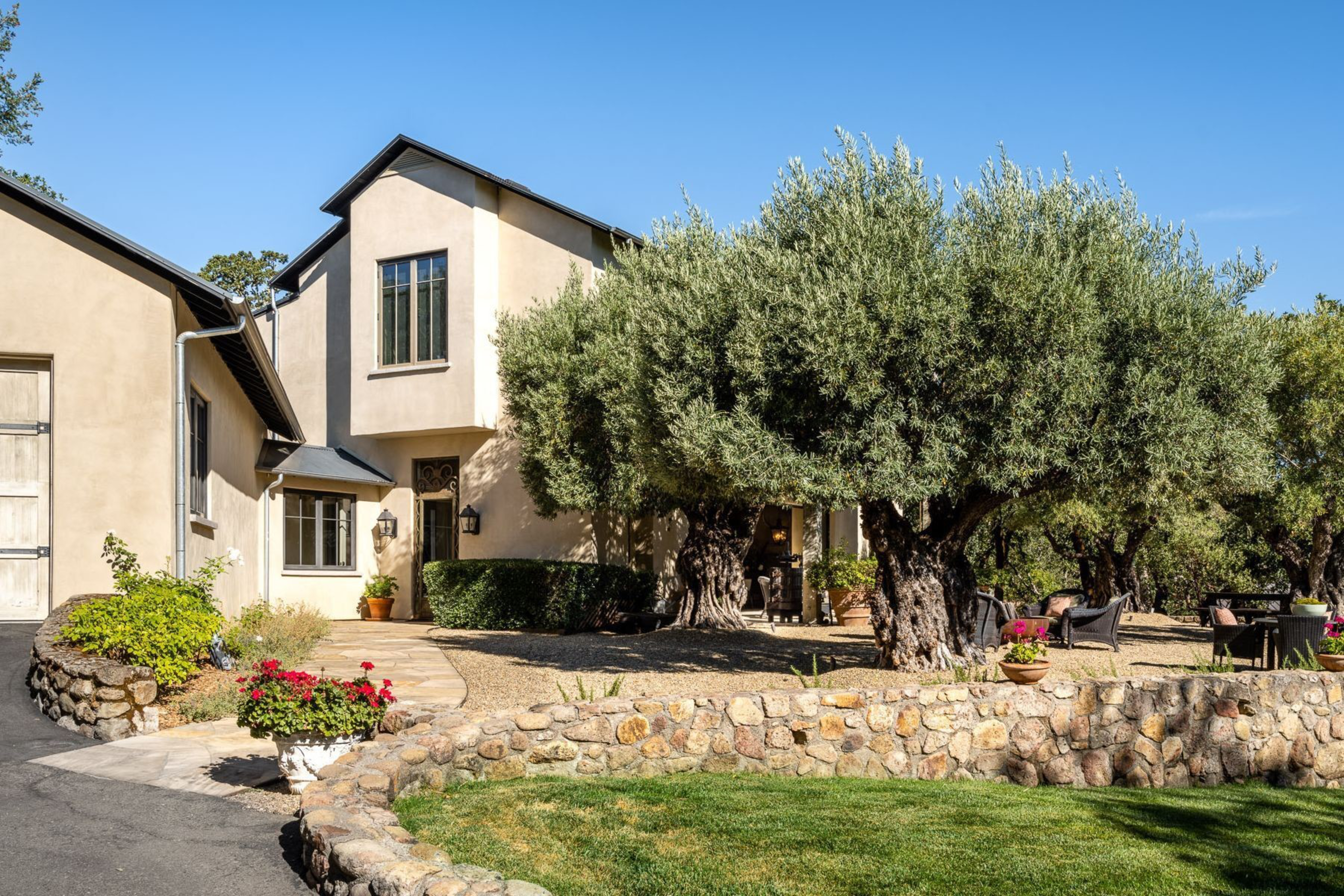A beige two-story house with large olive trees, a stone retaining wall, and outdoor seating with flower pots under clear blue skies.