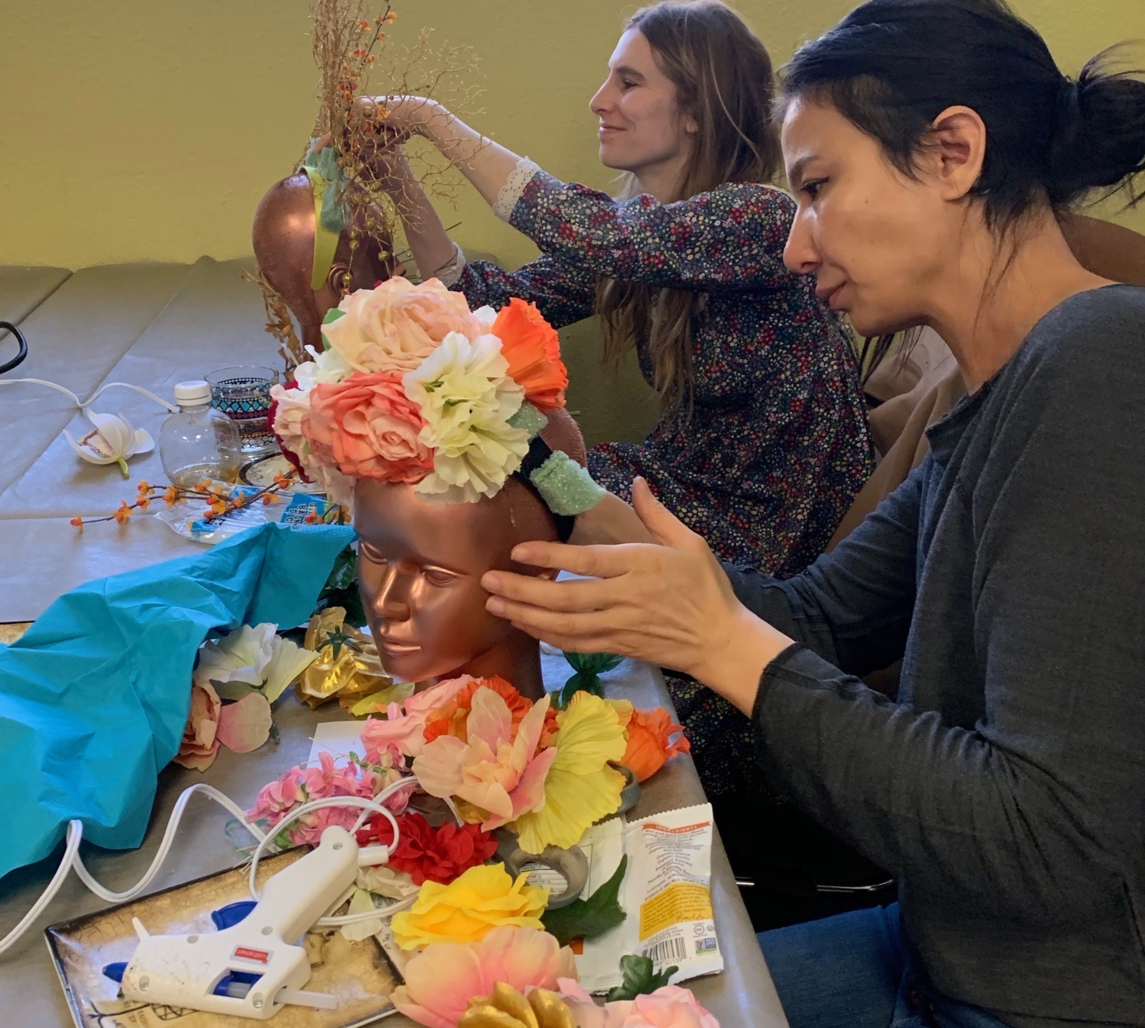 Two women are crafting floral headpieces on bronze mannequin heads, with various artificial flowers and a glue gun scattered on the table.