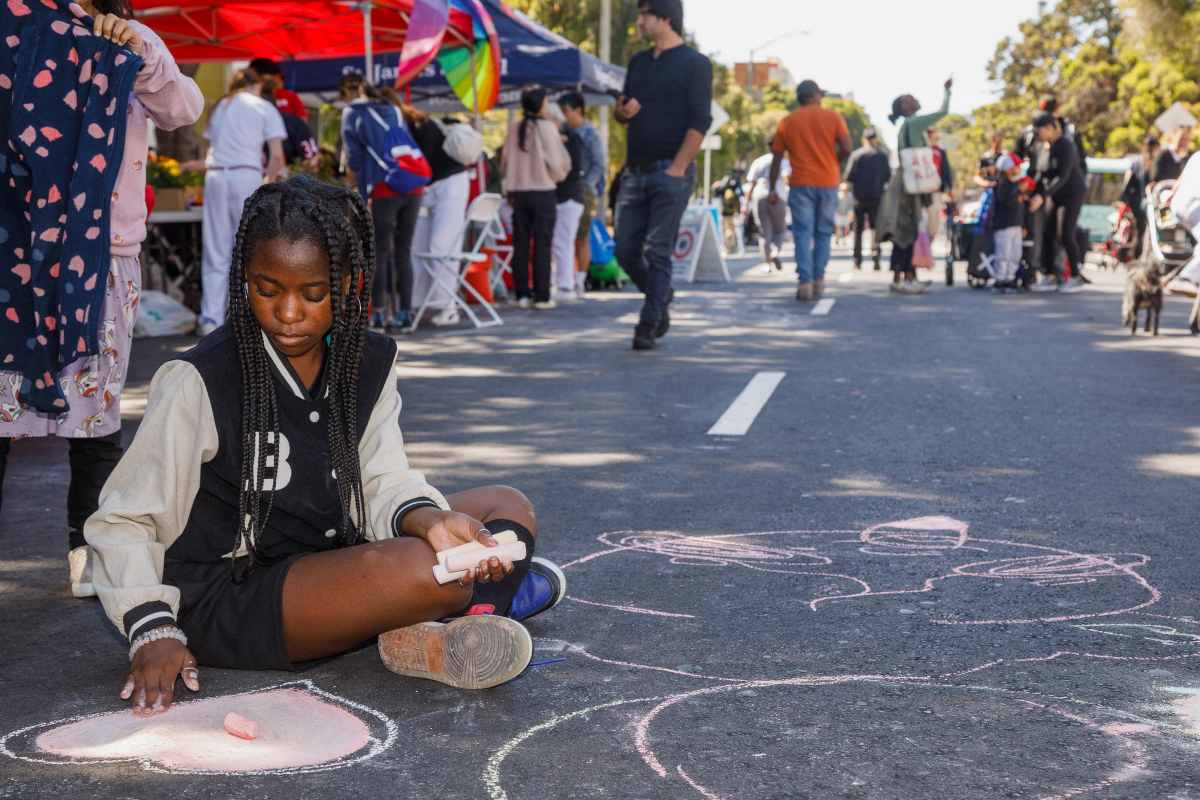 A young girl sits on a street drawing with chalk, with people and a market in the background.