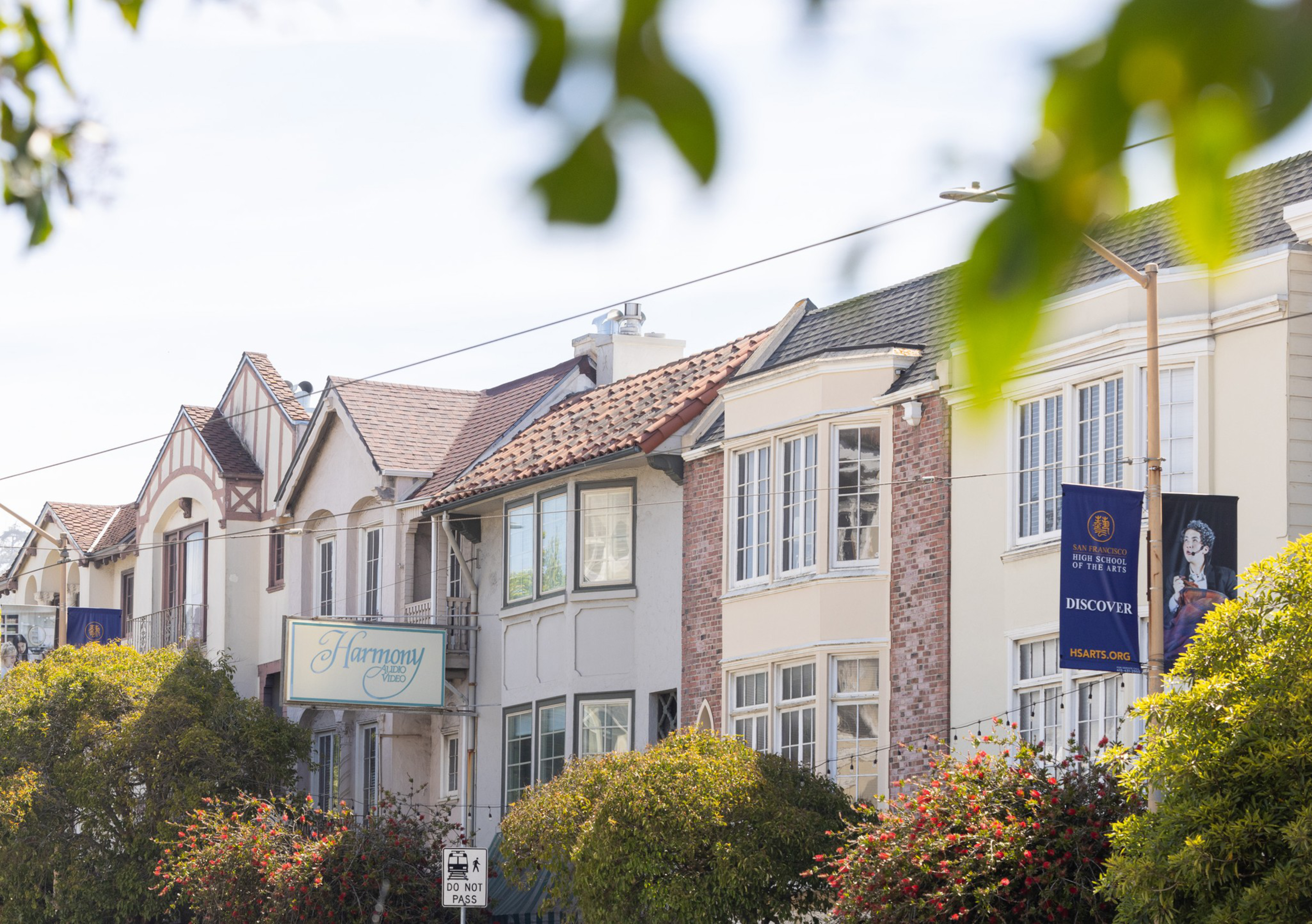 The image shows a row of charming, multi-story houses with various architectural styles, adorned with red-tiled roofs. Greenery and a banner for a high school are visible.