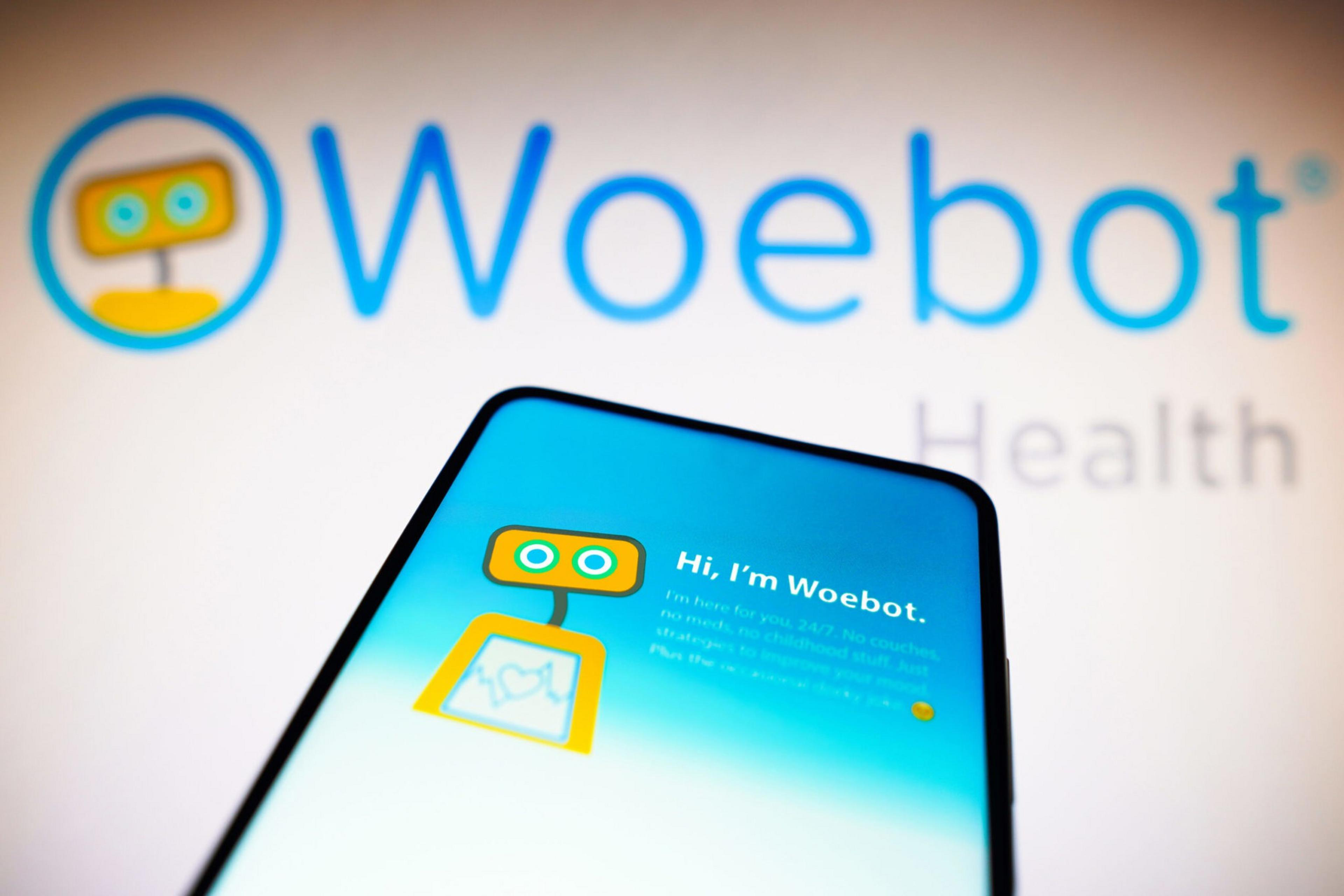 A smartphone screen displays a yellow and orange robot with the text "Hi, I'm Woebot" against a blurred background with the Woebot Health logo.