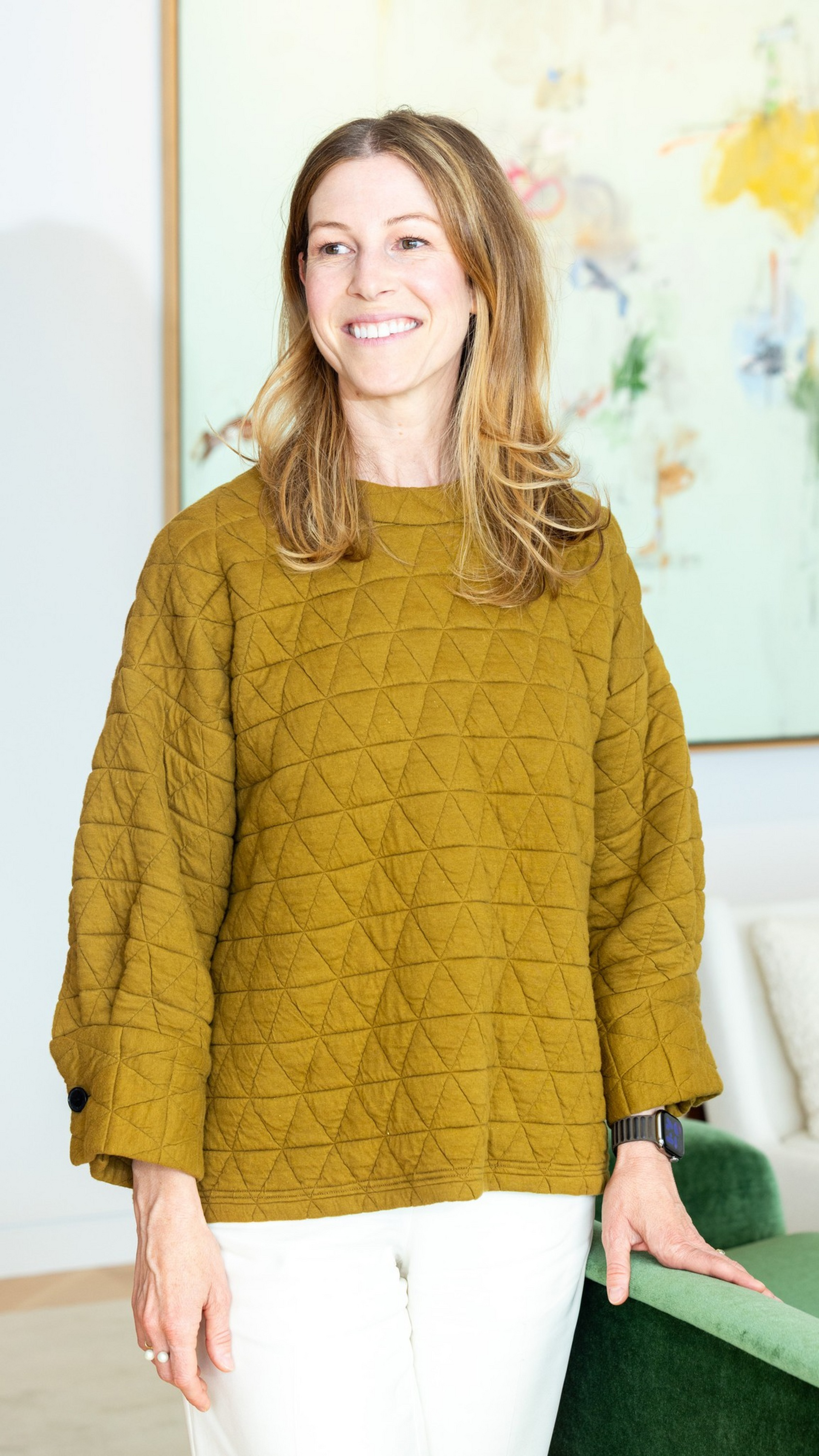 A smiling woman with light brown hair wears a mustard yellow quilted sweater and white pants, standing indoors near a green chair.