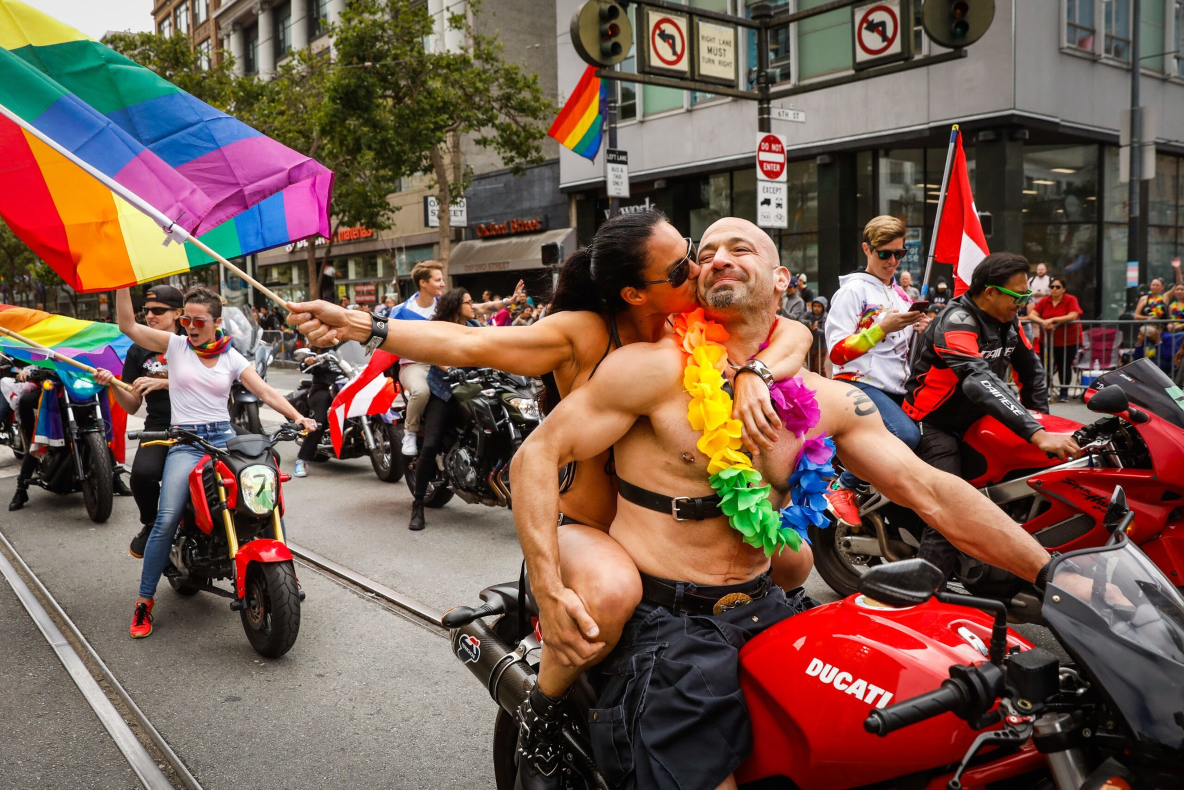 A muscular man on a red Ducati motorcycle wears rainbow leis while a woman hugs and kisses him, and a person nearby waves a large rainbow pride flag.