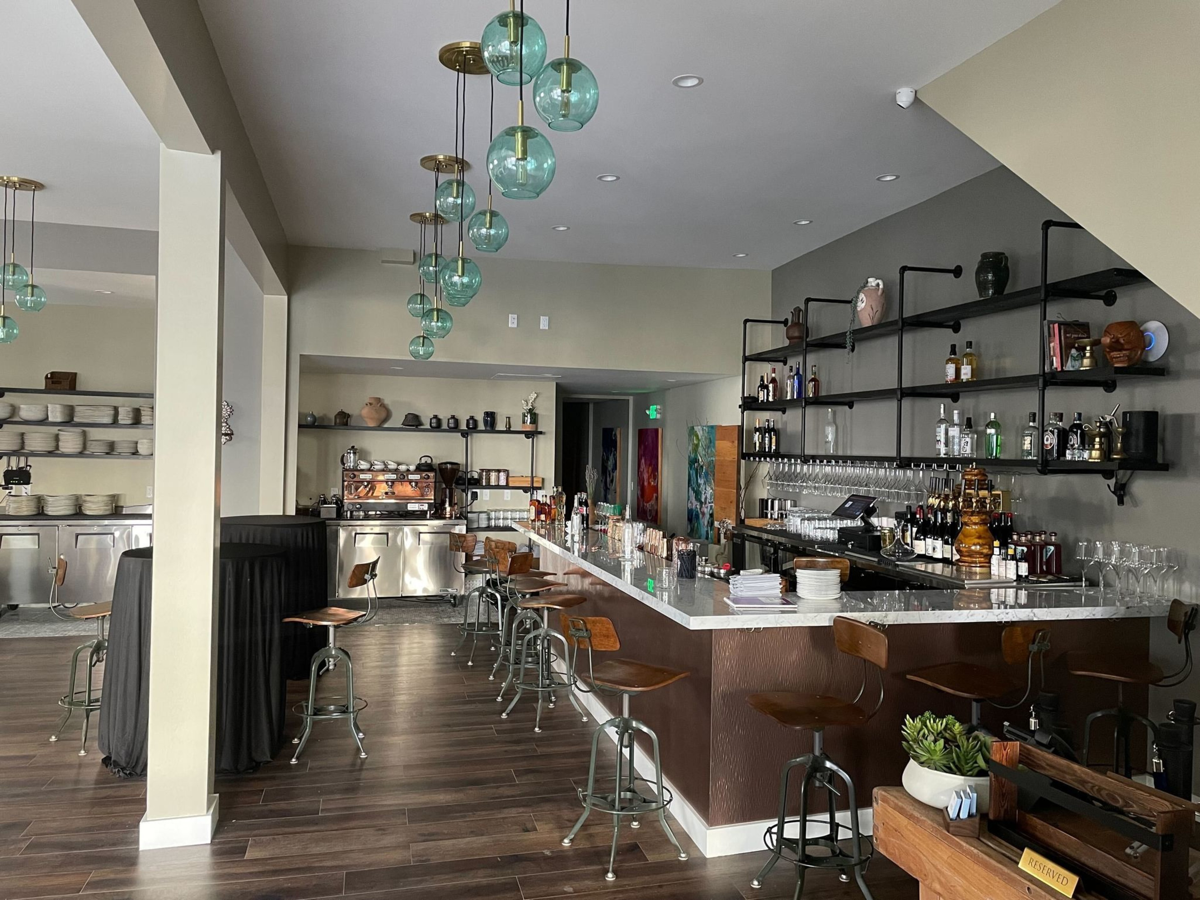 The image shows a modern bar with a long marble counter, high wooden stools, hanging glass bubble lights, and shelves stocked with bottles and glasses.