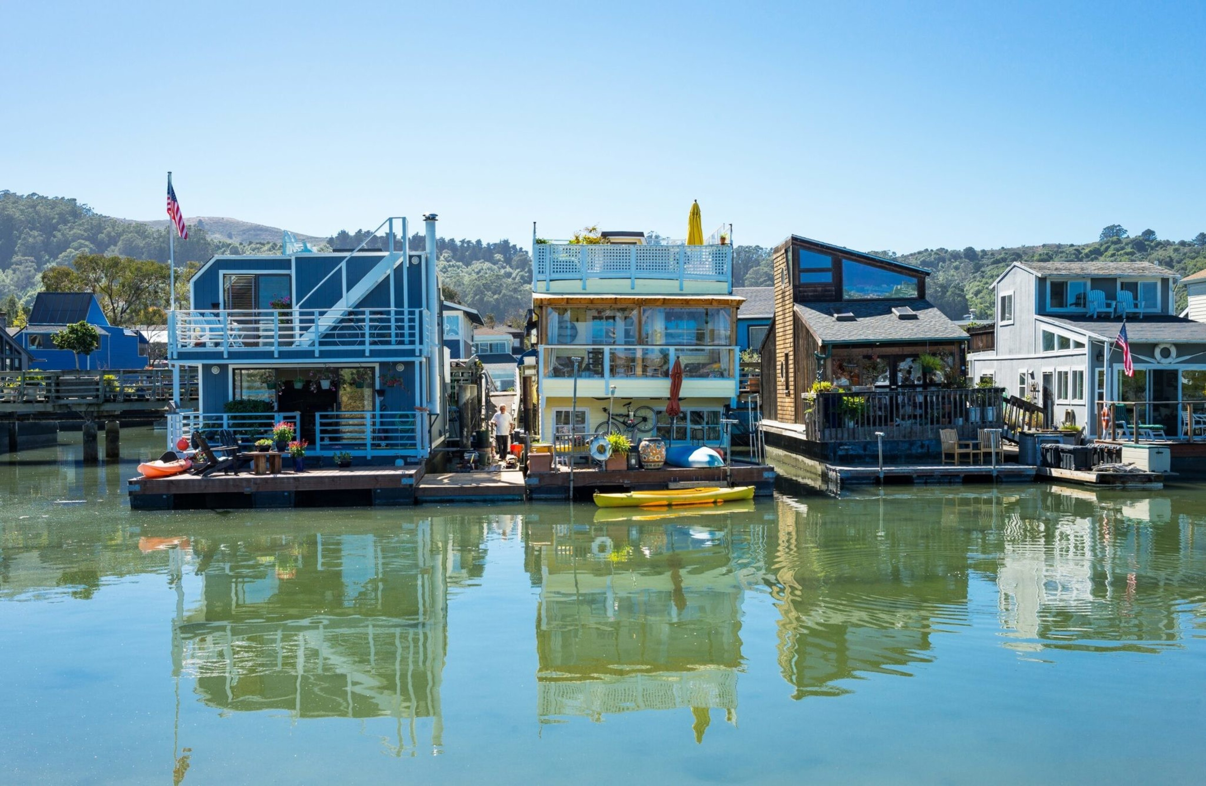 A row of colorful houseboats floats peacefully on calm water, each uniquely designed, with decks adorned by plants and outdoor furniture under clear skies.