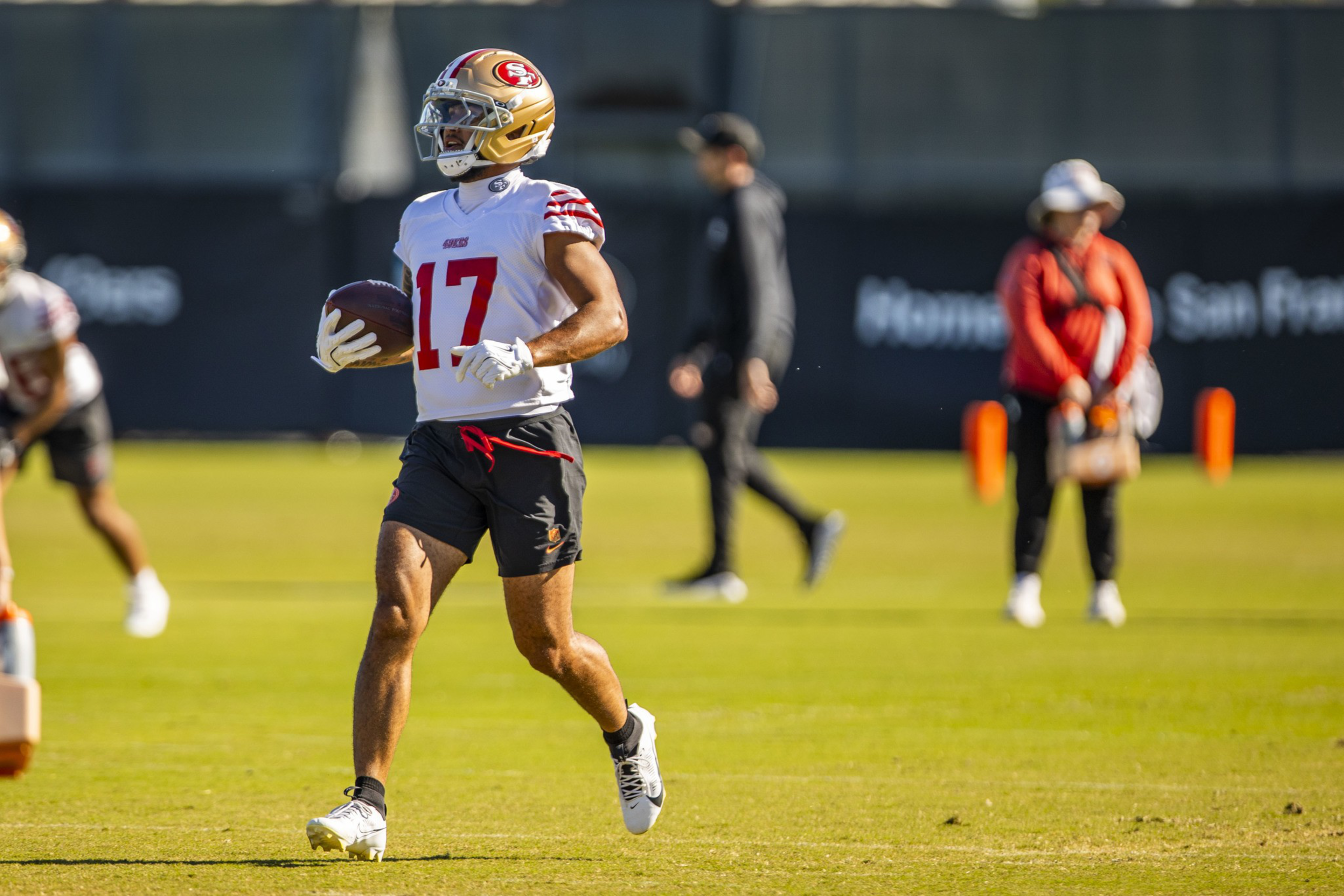 A football player in a San Francisco 49ers helmet and jersey number 17 is running on a grass field holding a football.