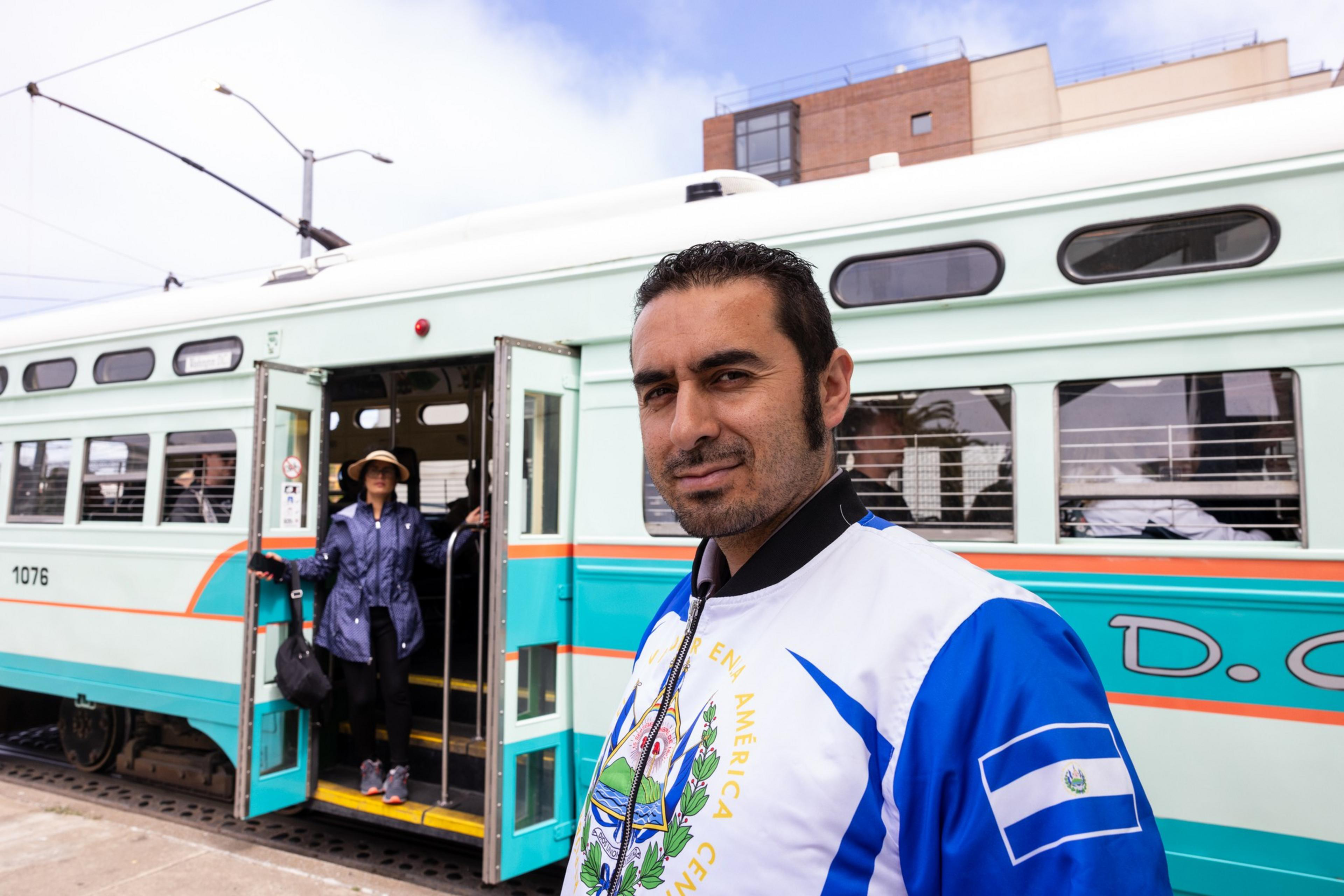 A man in a jacket with a flag stands in front of a light green and white trolley with open doors, while a woman in a hat exits behind him.