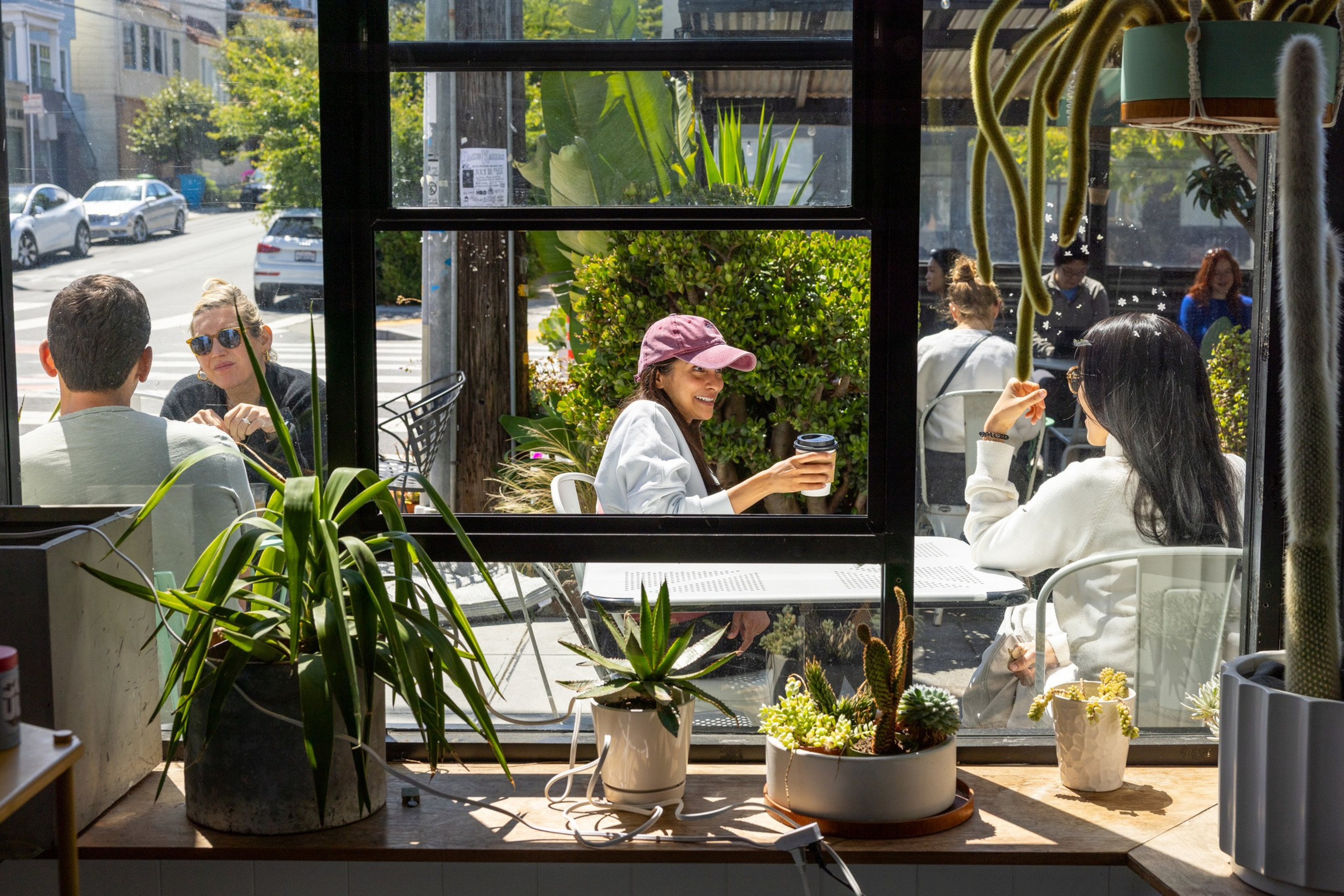 Two women chat and drink coffee outside a café window, surrounded by green plants and bright sunlight on a city street.