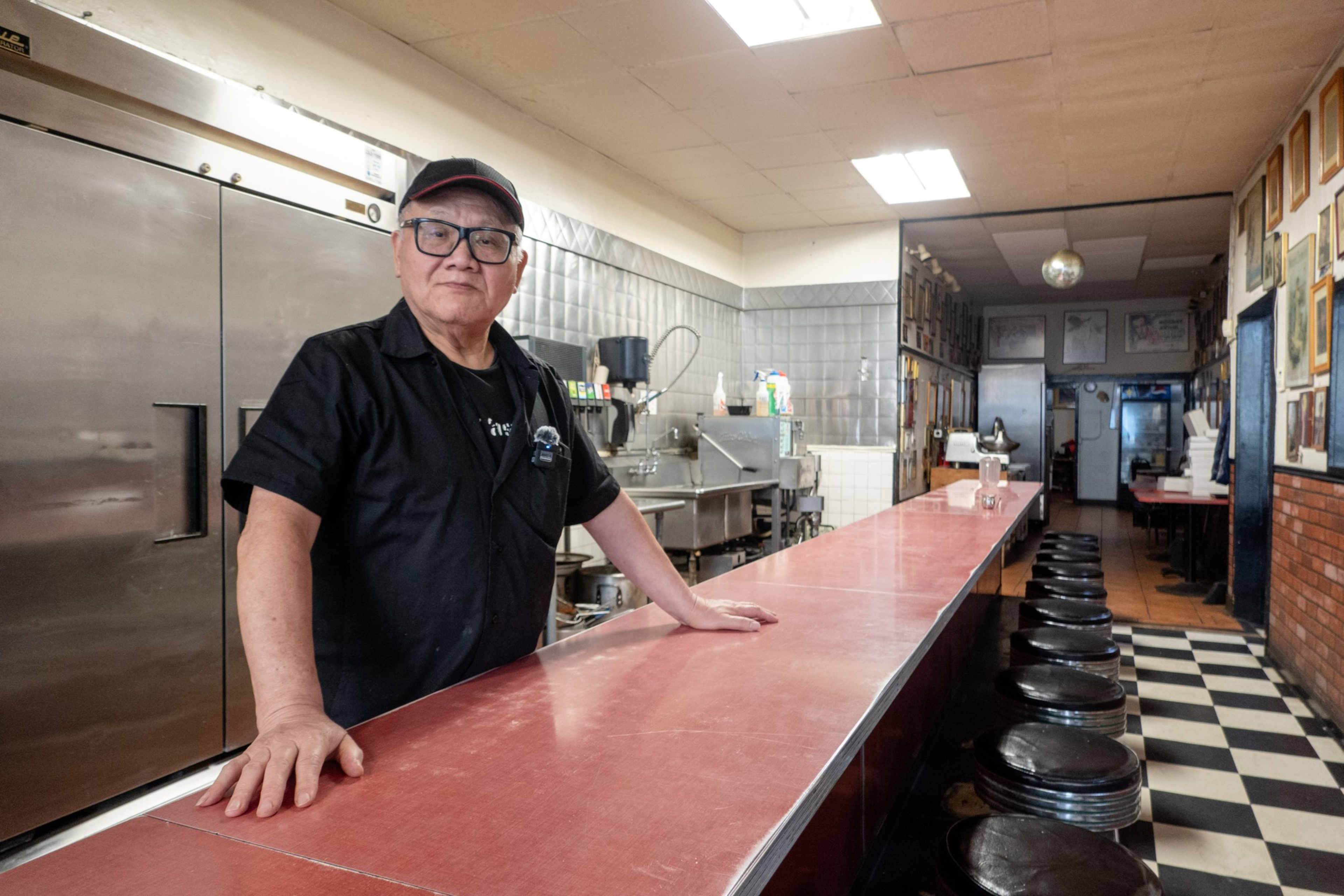 A person stands behind a diner counter with stools, walls adorned with pictures, and checkered floors.