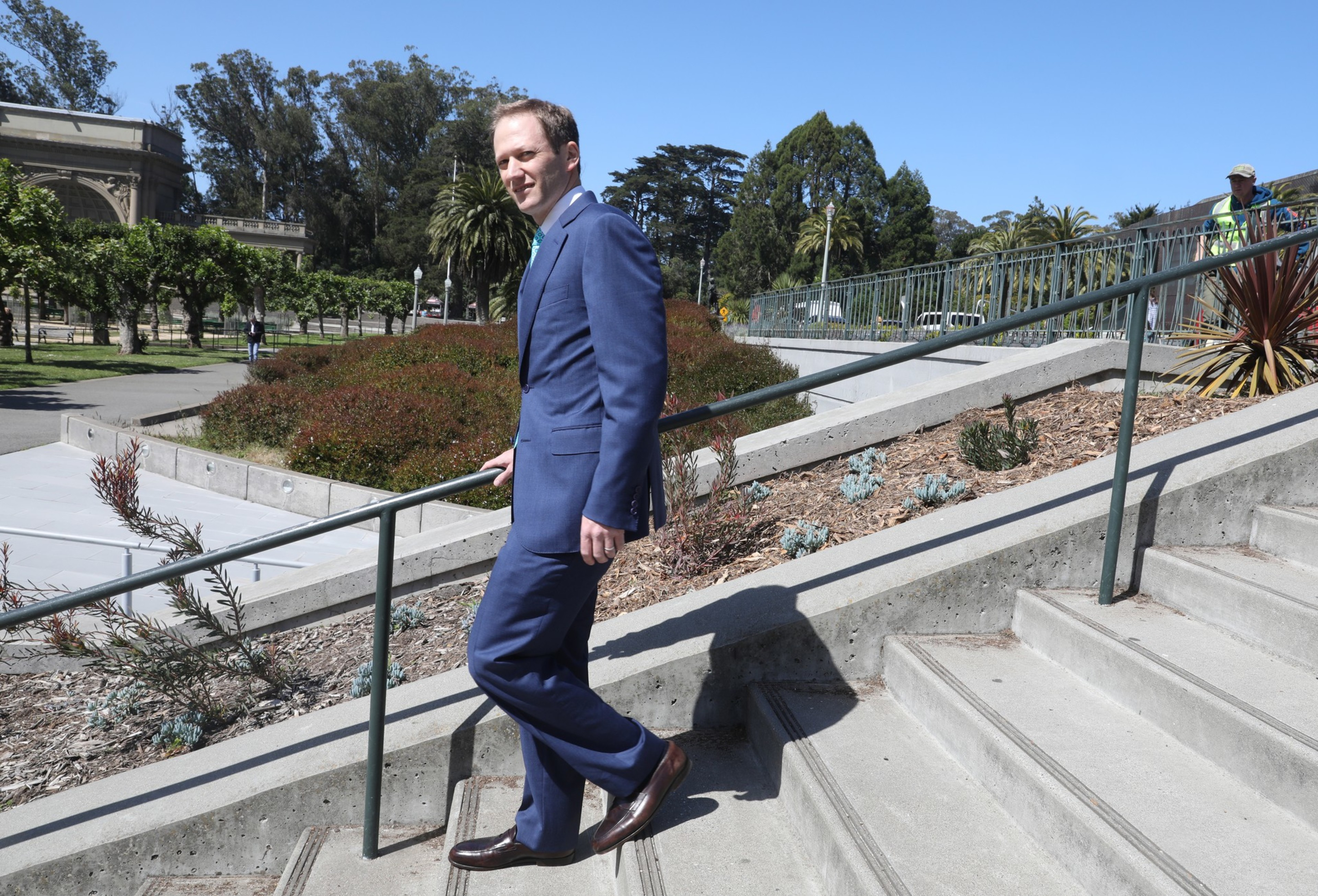 A man in a blue suit walks up concrete steps with greenery and trees around. The sky is clear and blue, and a railing follows the stairs.