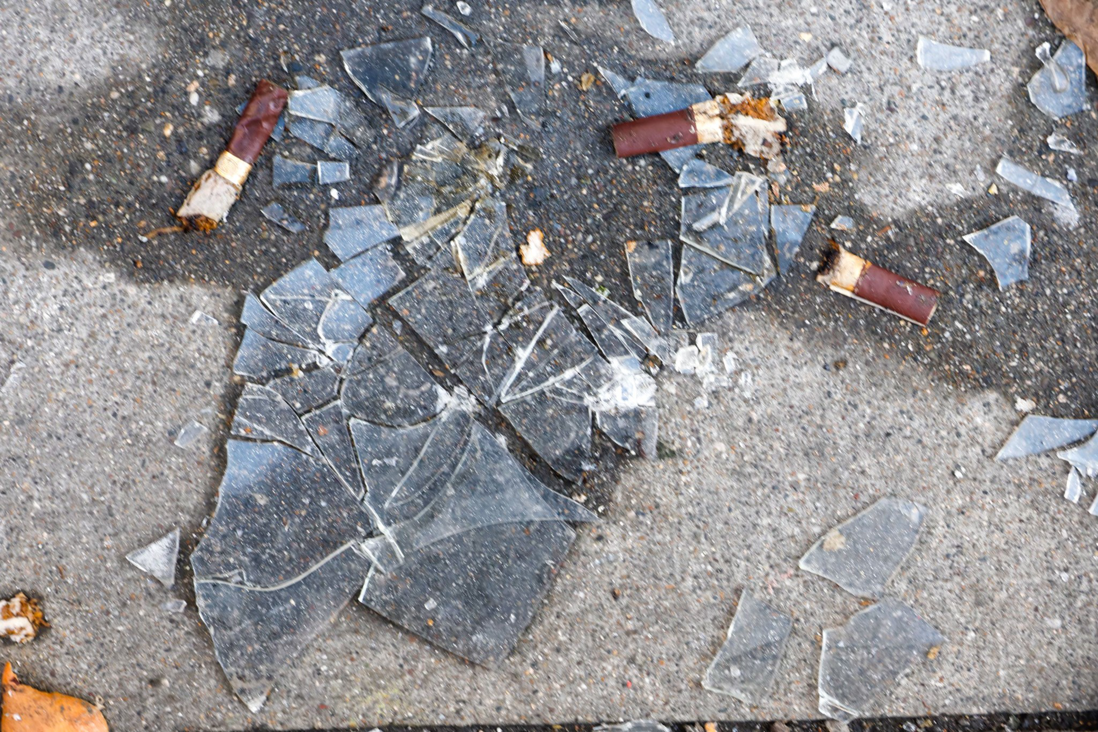 Broken glass shards are scattered on the ground alongside several cigarette butts on a concrete surface.