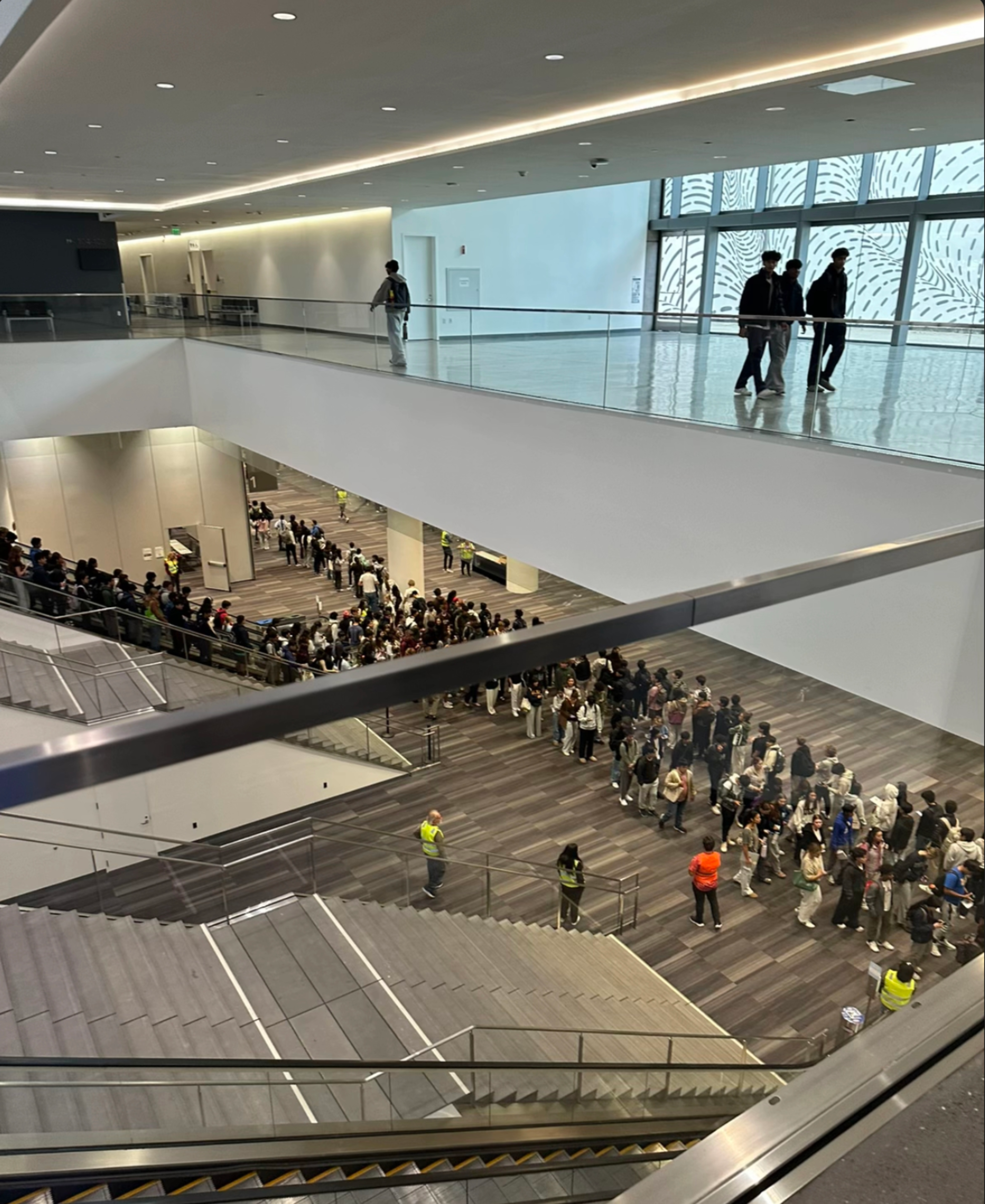 A large crowd forms a winding line inside a modern building with multiple floors and glass railings, while a few people stand on the upper level.