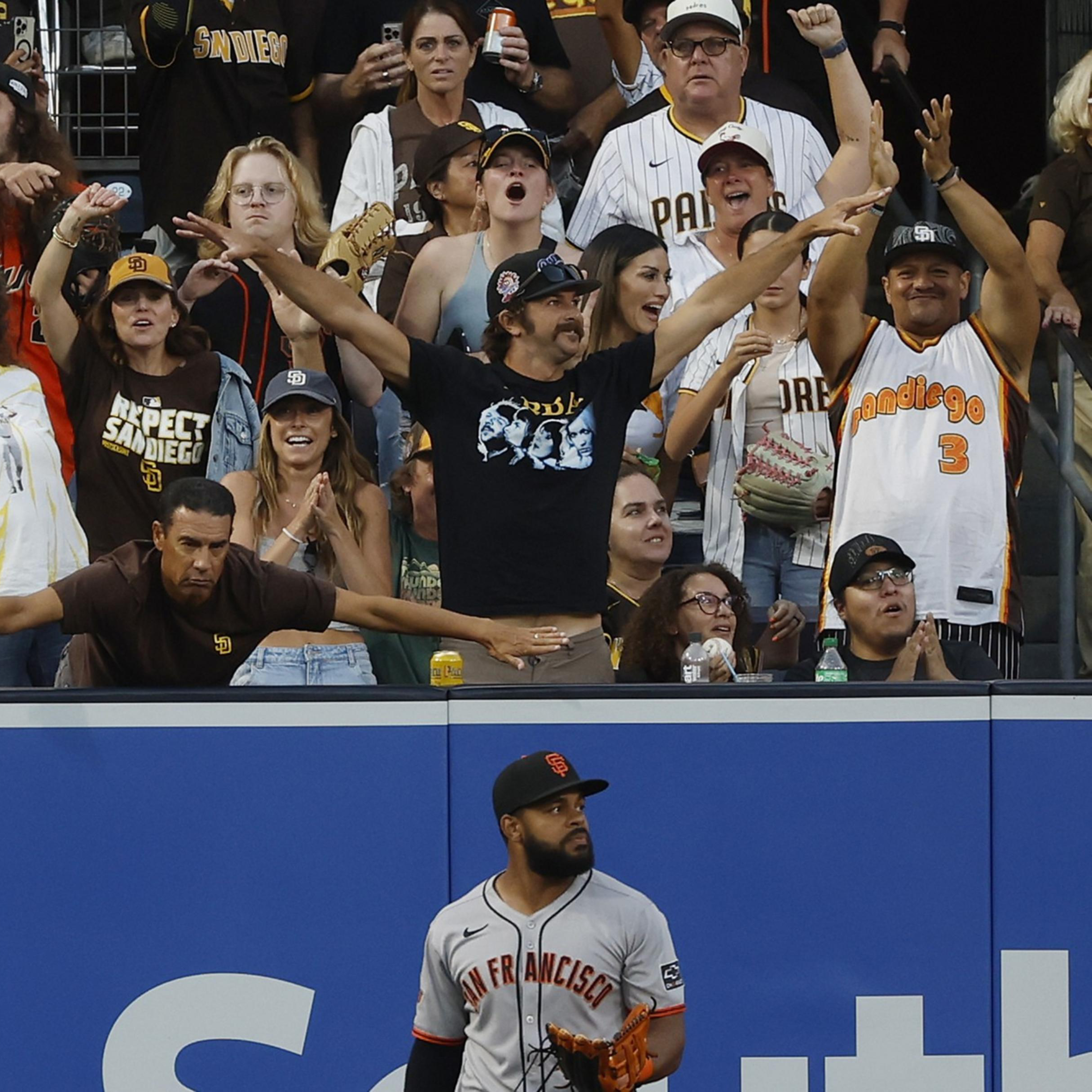 A San Francisco Giants player stands near the outfield wall as energetic fans behind him, mostly wearing San Diego Padres gear, cheer and react.
